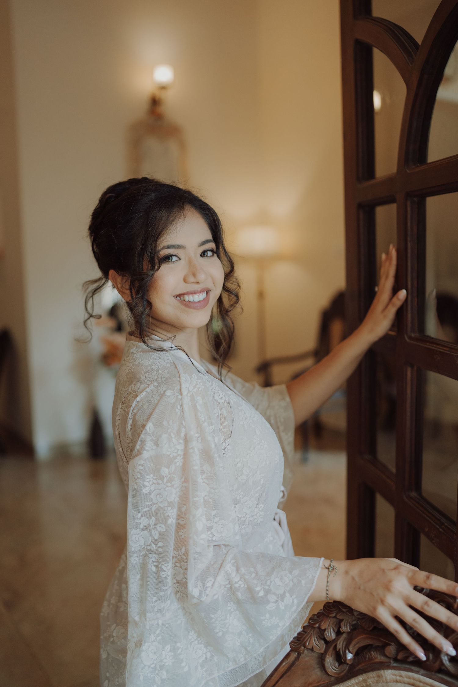 A smiling bride in a lace robe with an elegant updo leans against a wooden glass-paneled door while getting ready for her wedding.