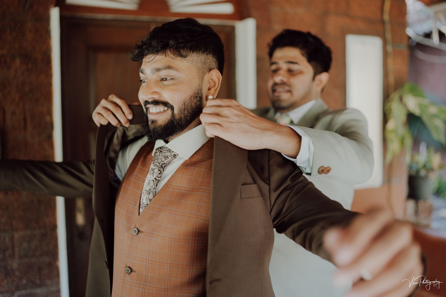 A groomsman helps adjust the groom's brown suit vest during wedding preparations at an indoor venue.