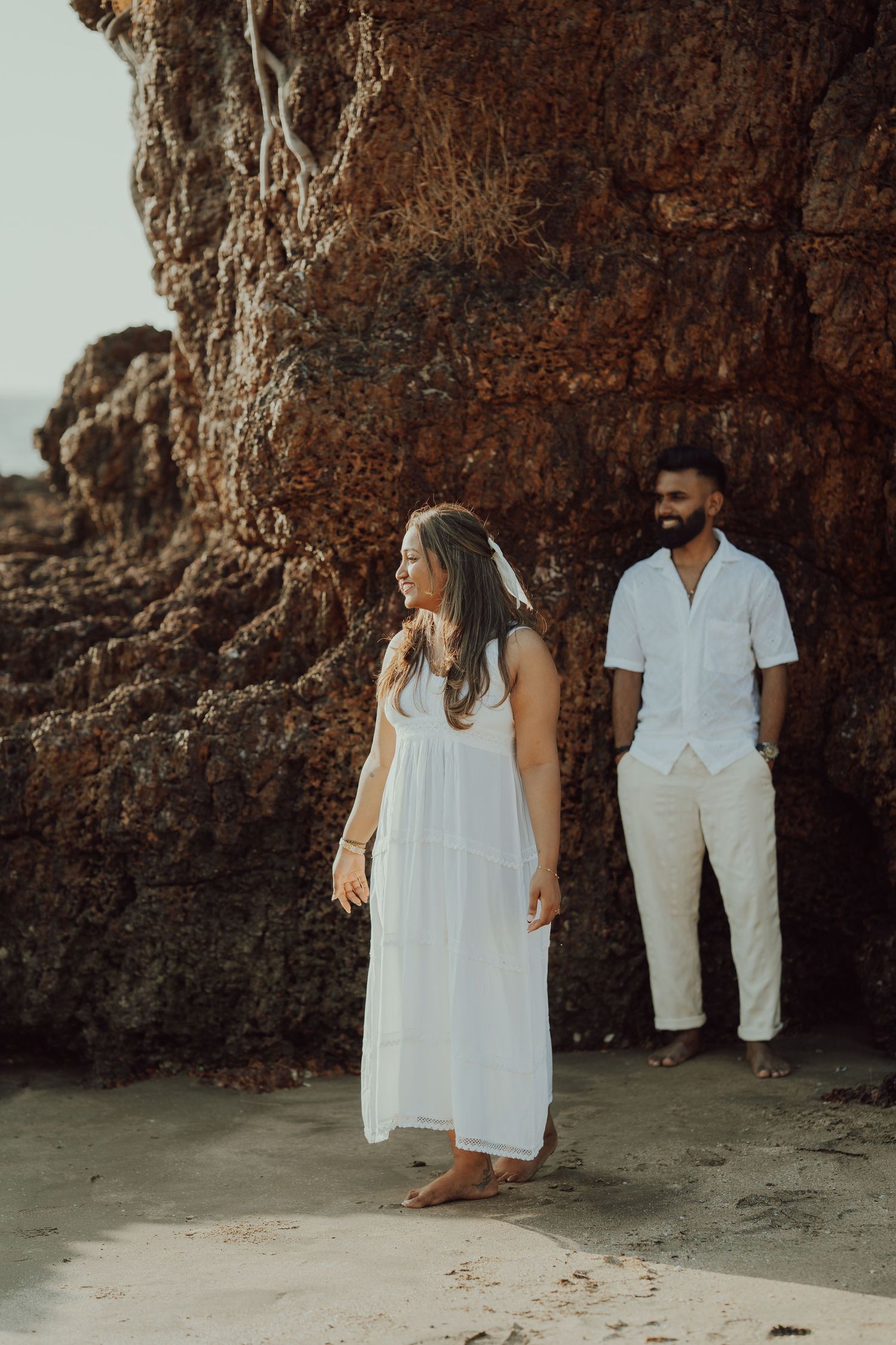 A couple in white outfits walking along a rocky beach cave during sunset, creating romantic silhouettes against the cliffs.