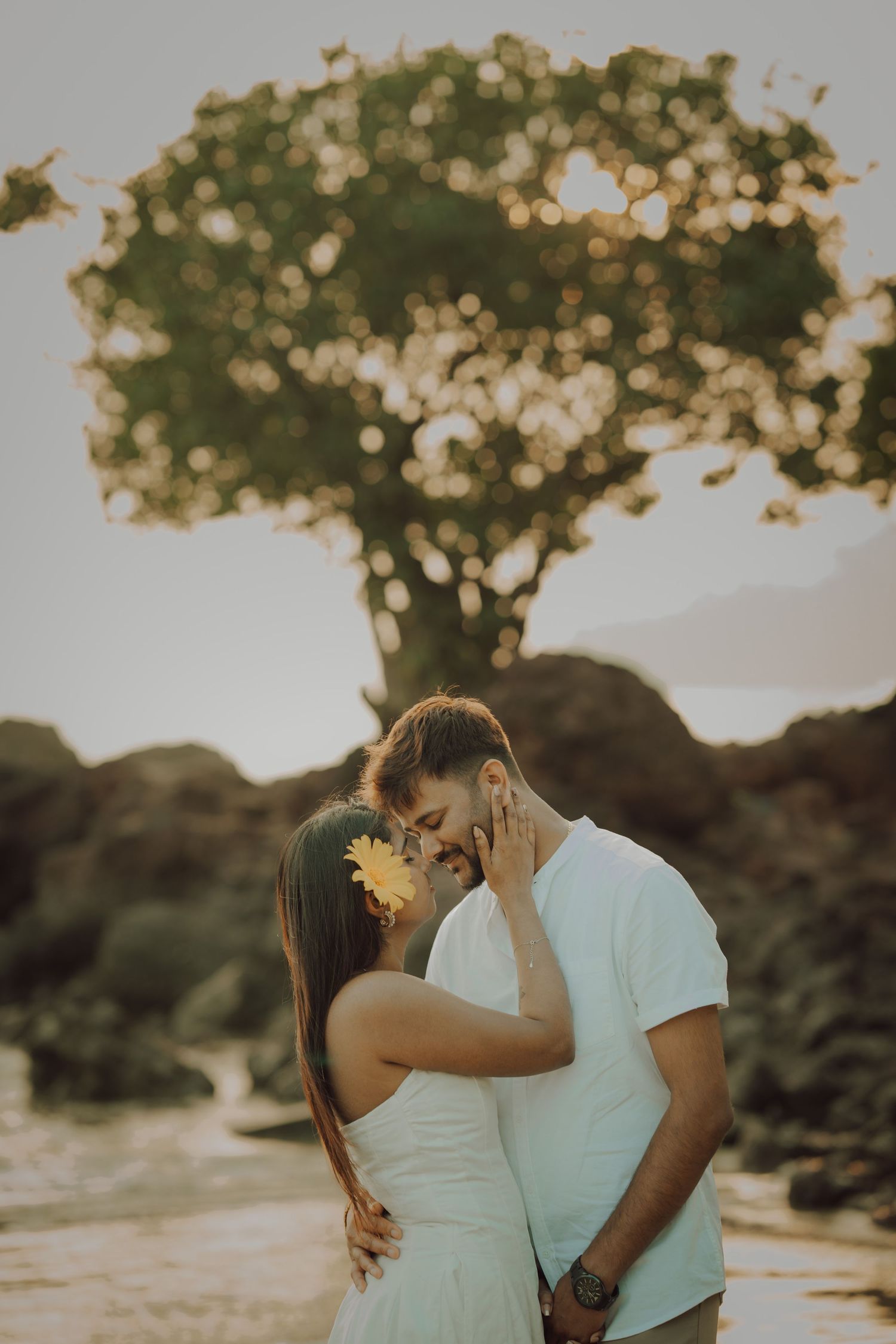 Romantic couple embraces and kisses at sunset in a natural outdoor setting with trees in the background.