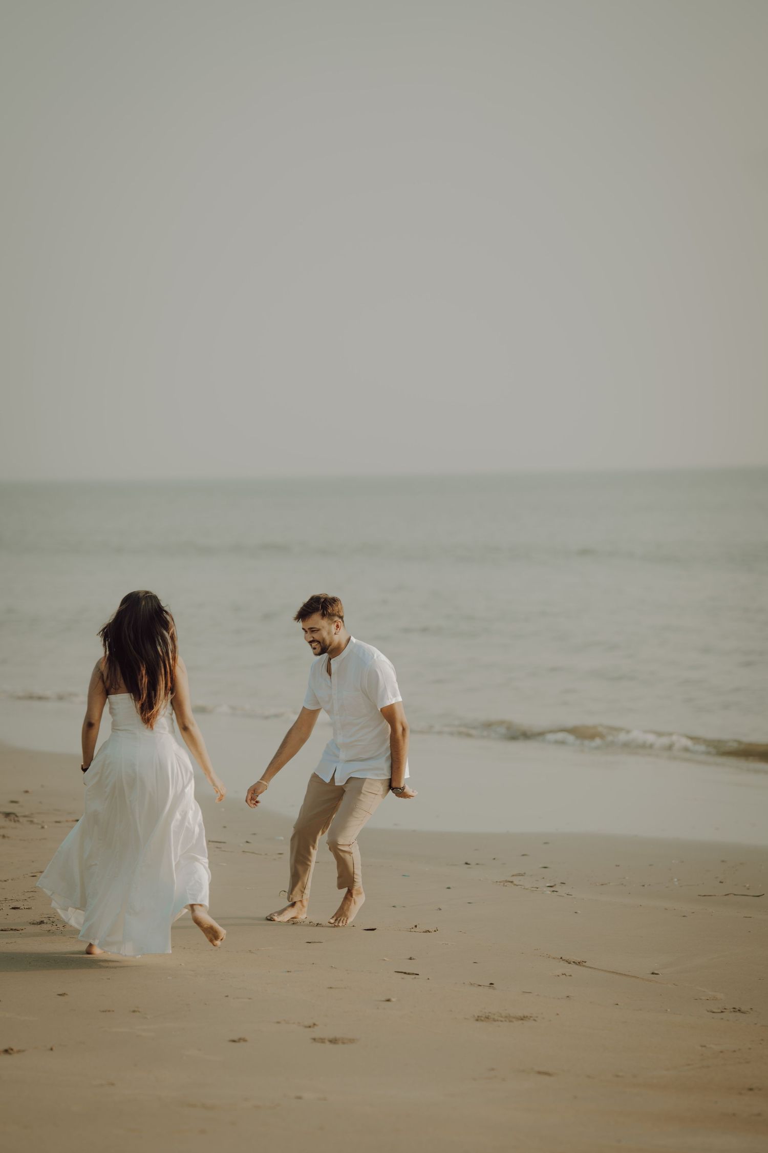 A couple in white and beige clothing searches for seashells together on a misty beach at sunset.
