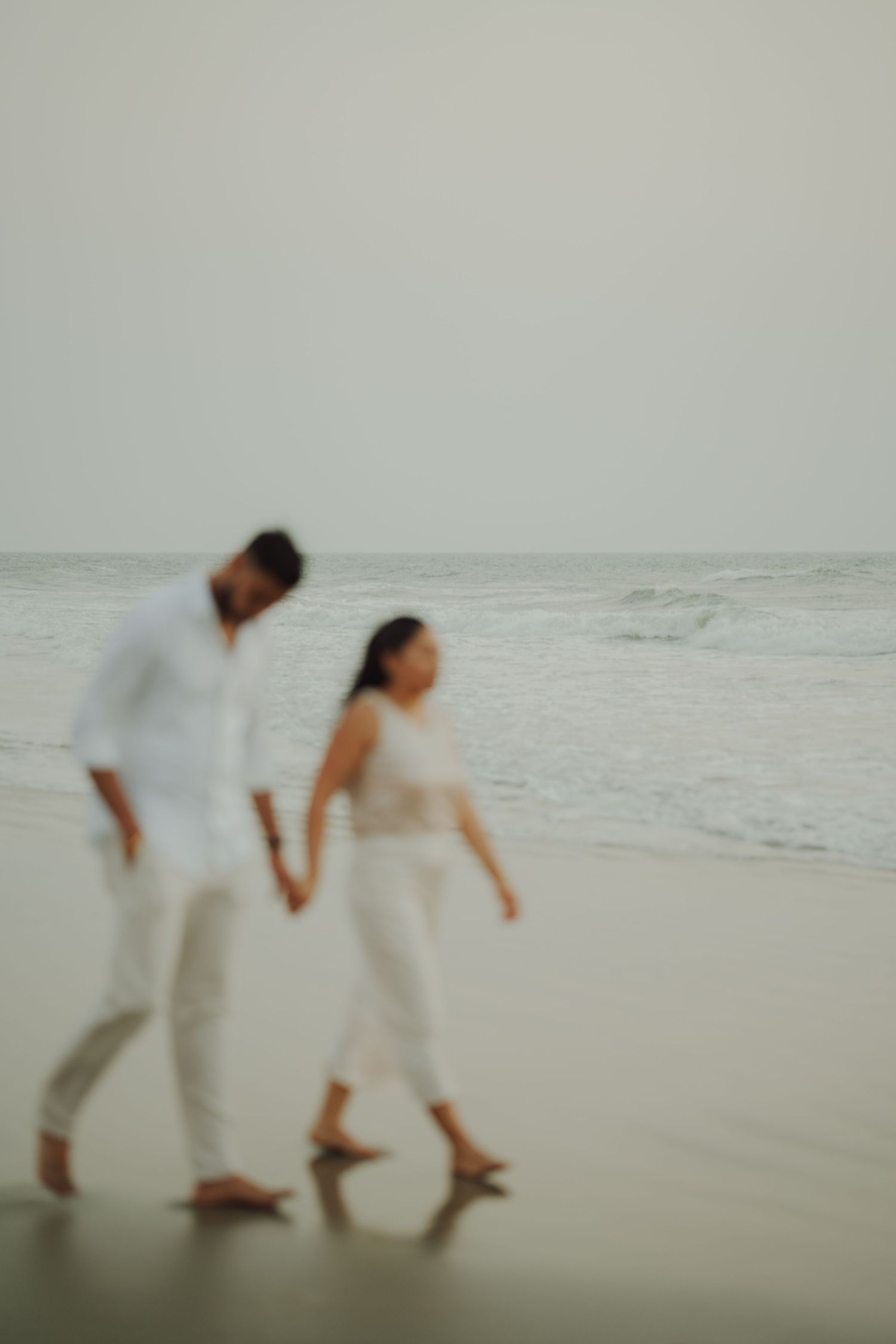 Couple holding hands walking barefoot on a beach in white outfits, with ocean waves in the background.