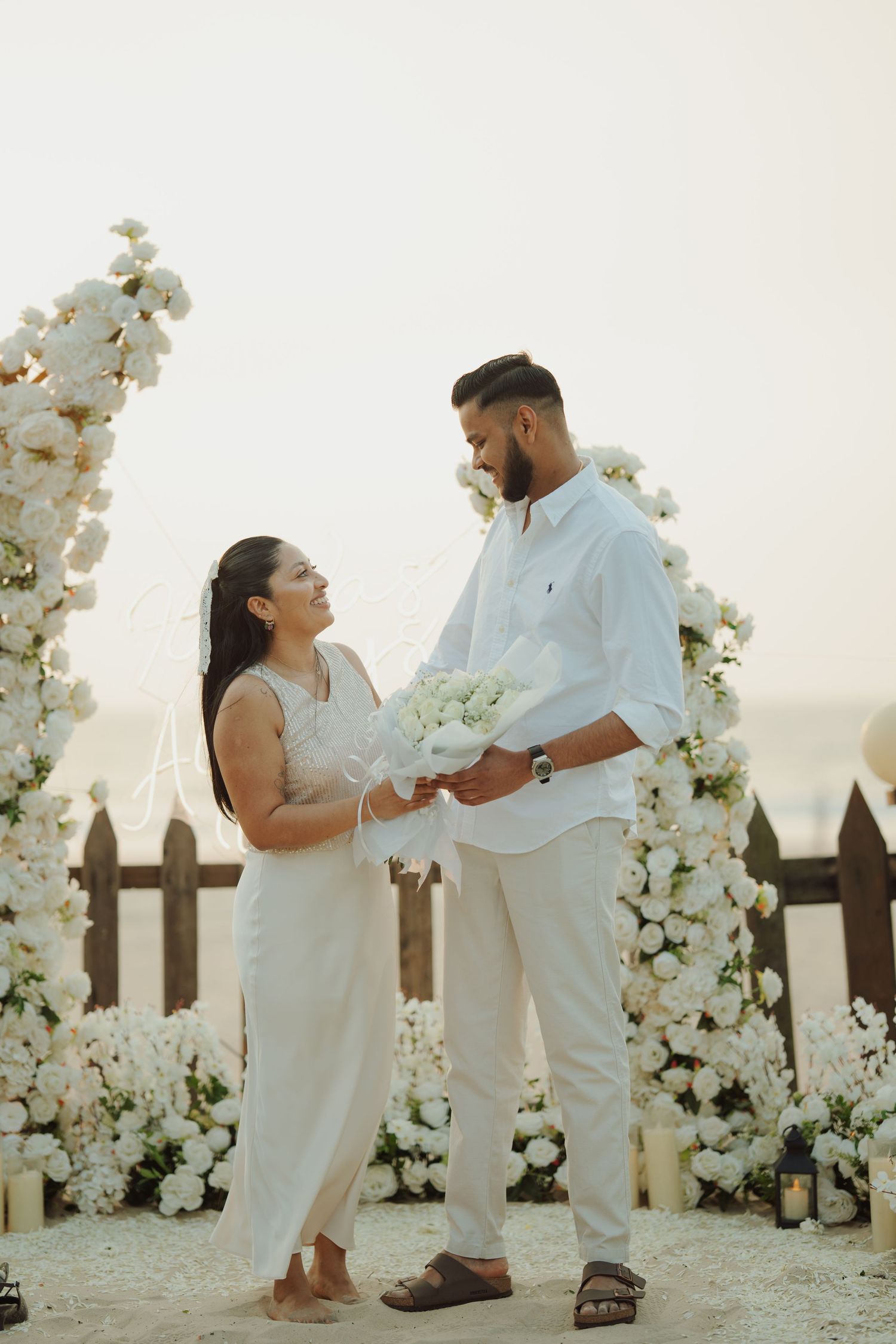 Couple in white attire exchanging vows surrounded by white floral arches on a beach at sunset.