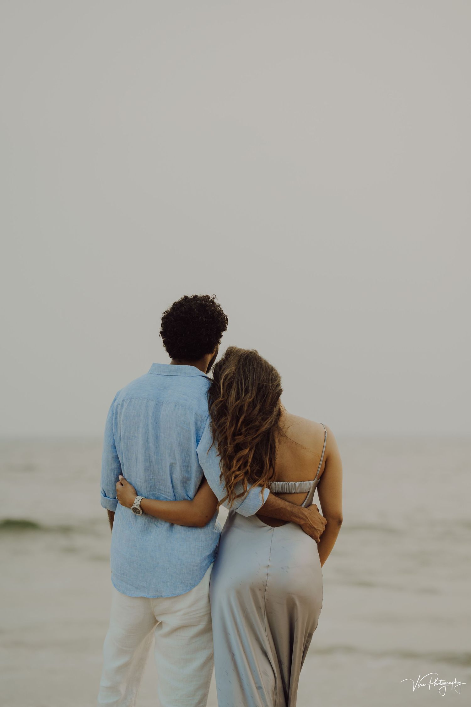 Romantic couple embraces while looking at ocean view, shot from behind on misty beach at dusk.