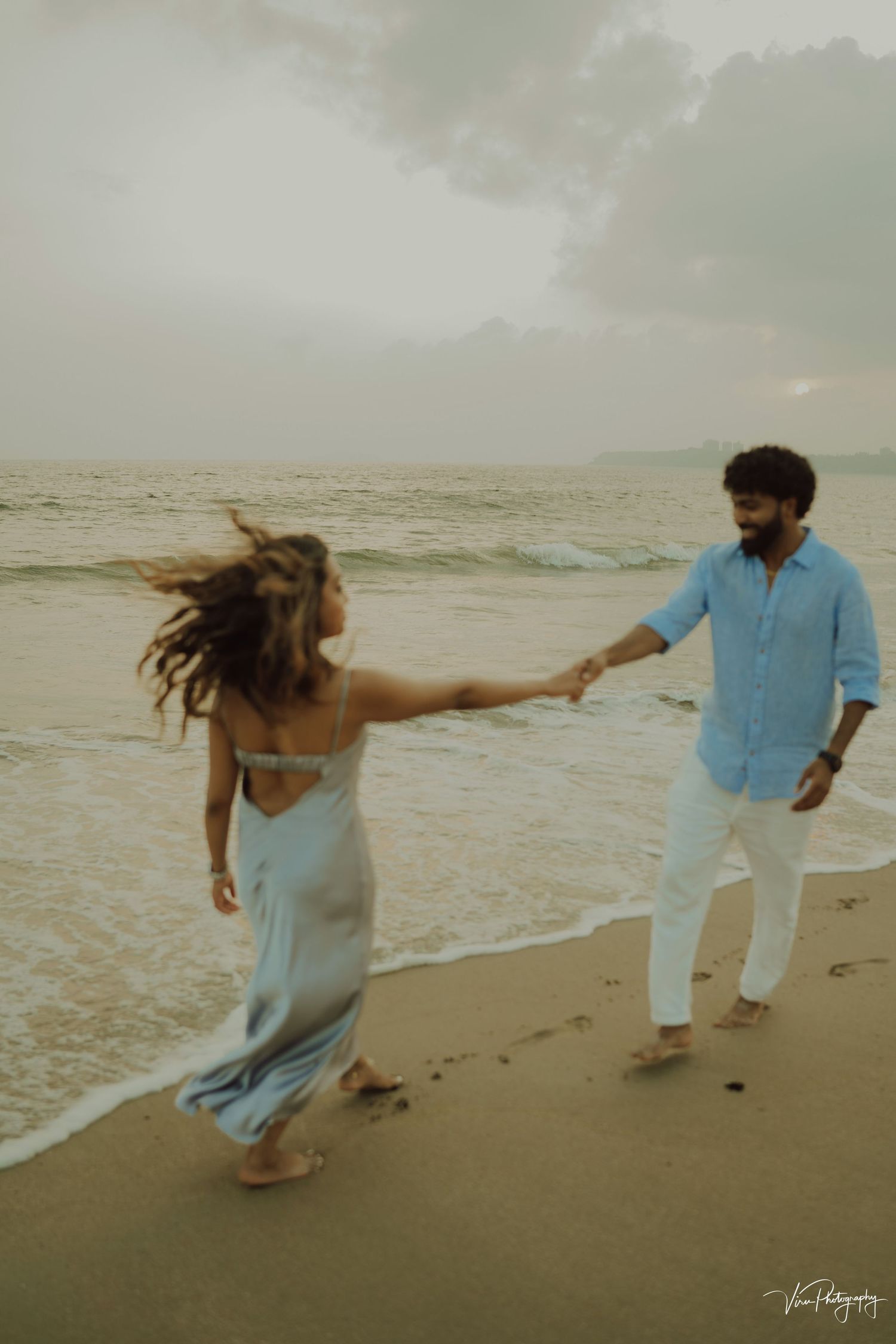 A couple holds hands and dances on a misty beach at sunset, with her dress flowing in the sea breeze.