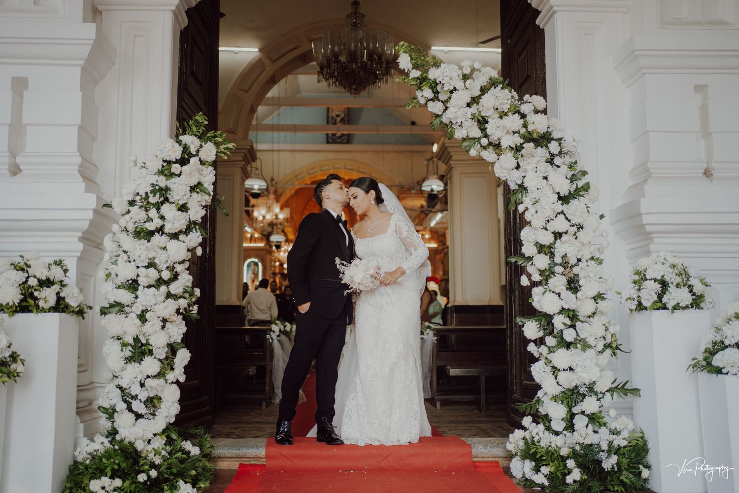 A romantic wedding ceremony setup with an elegant white floral arch and red carpet at a grand entrance.