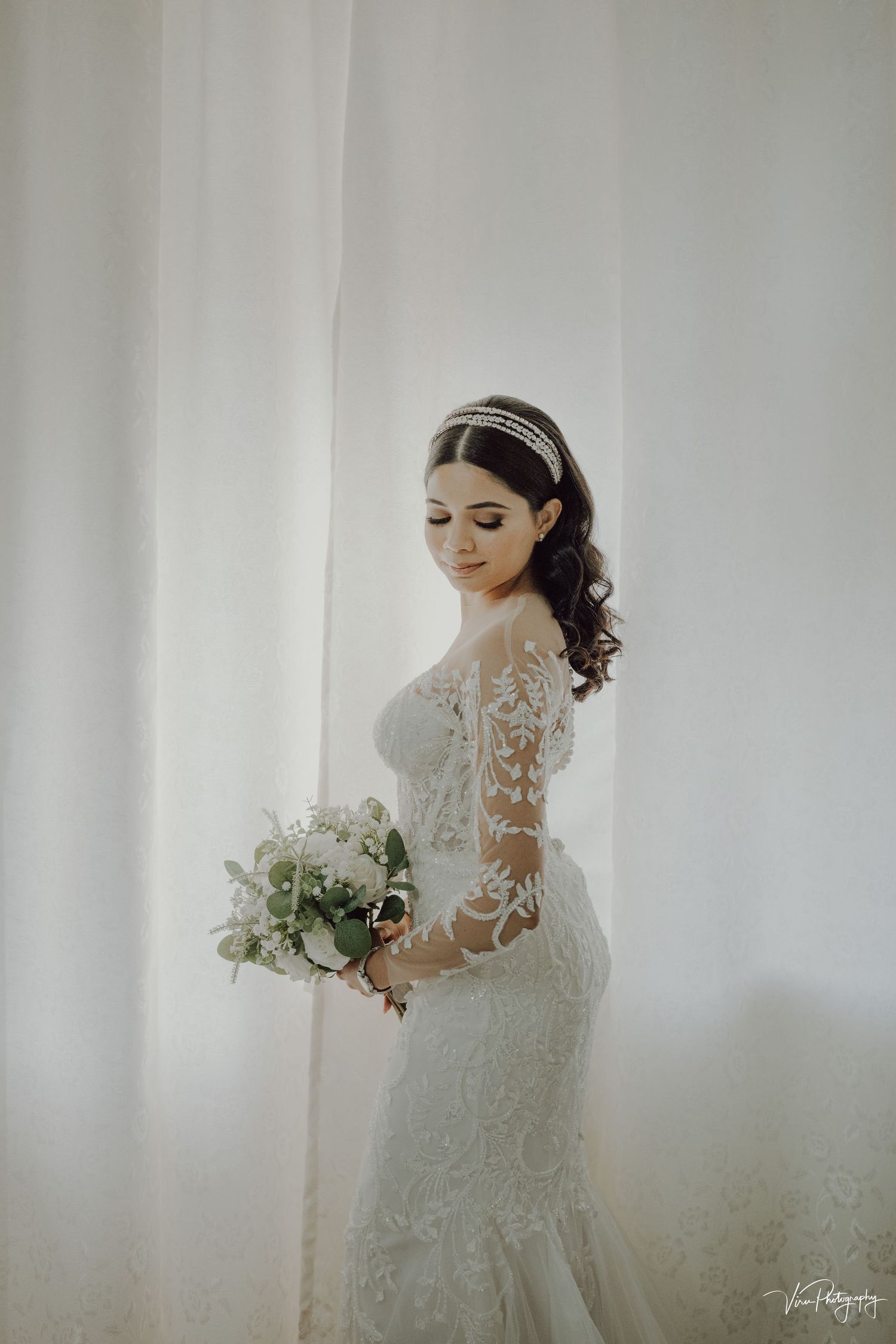 Ethereal bride in long-sleeved lace wedding gown holds white bouquet against light curtained background in soft lighting.