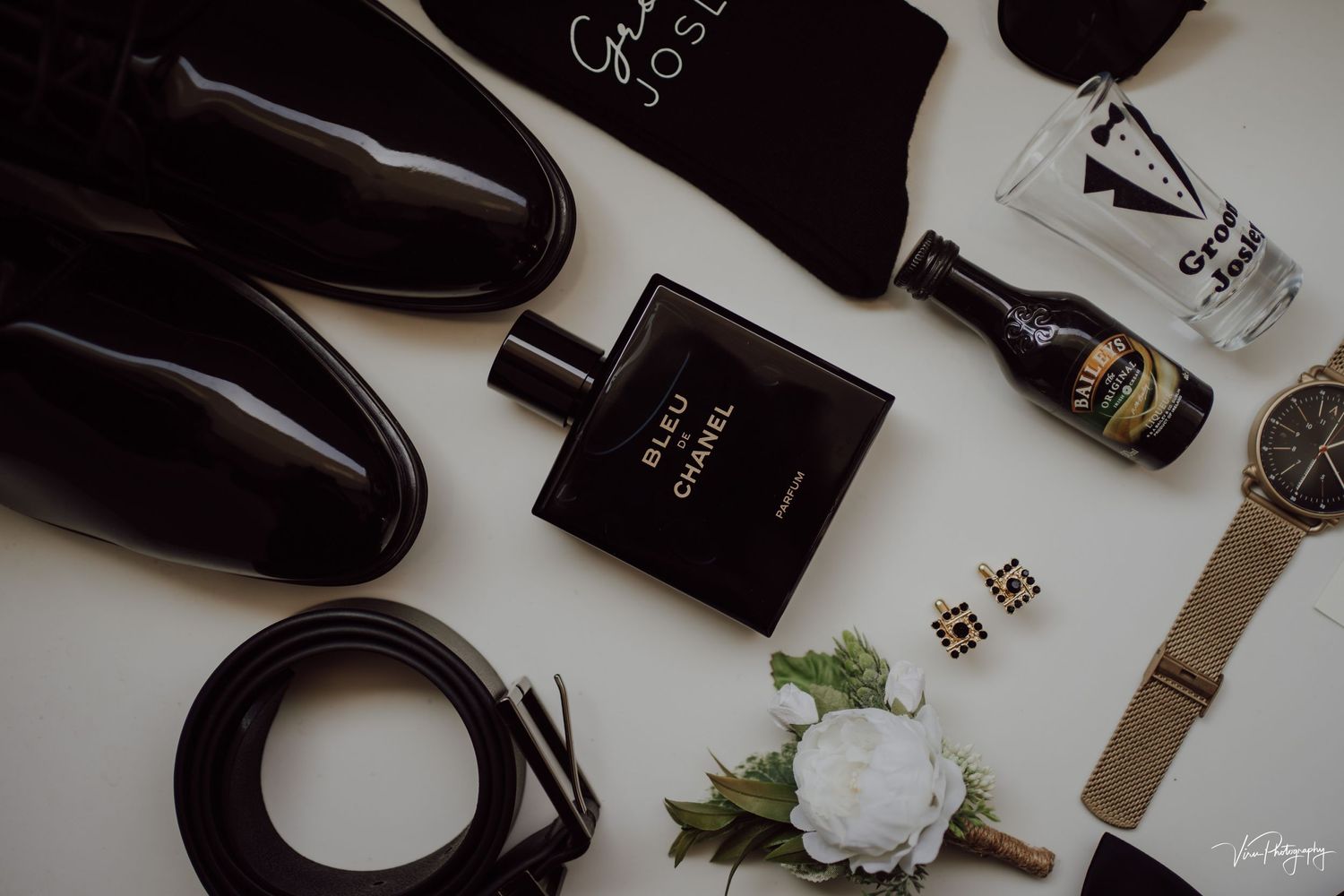 Luxury men's accessories including black leather shoes, cologne, wristwatch and flower arrangement on white surface.
