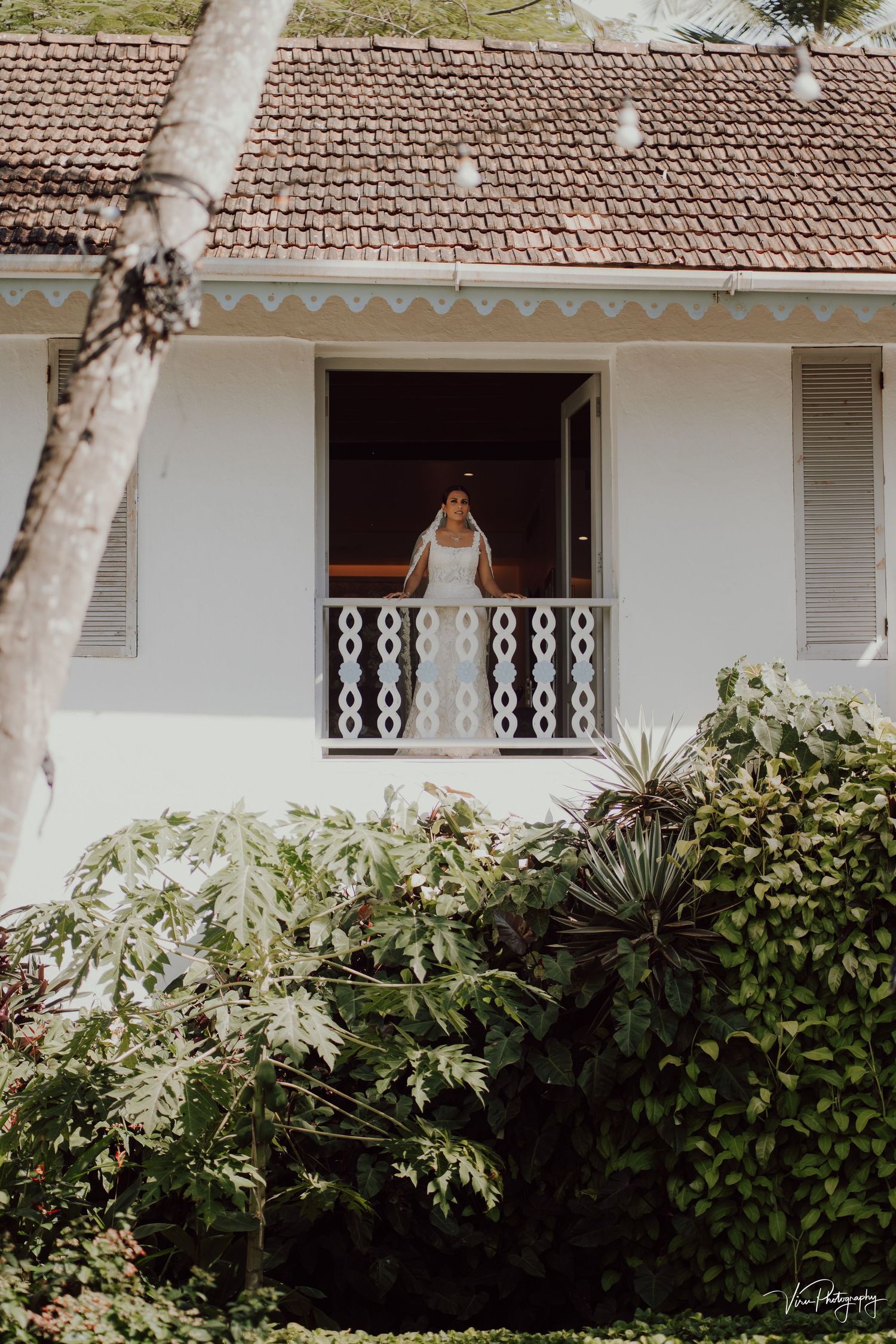 A wooden-framed doorway with decorative white balusters opens to a balcony on a white stucco house with tropical landscaping.