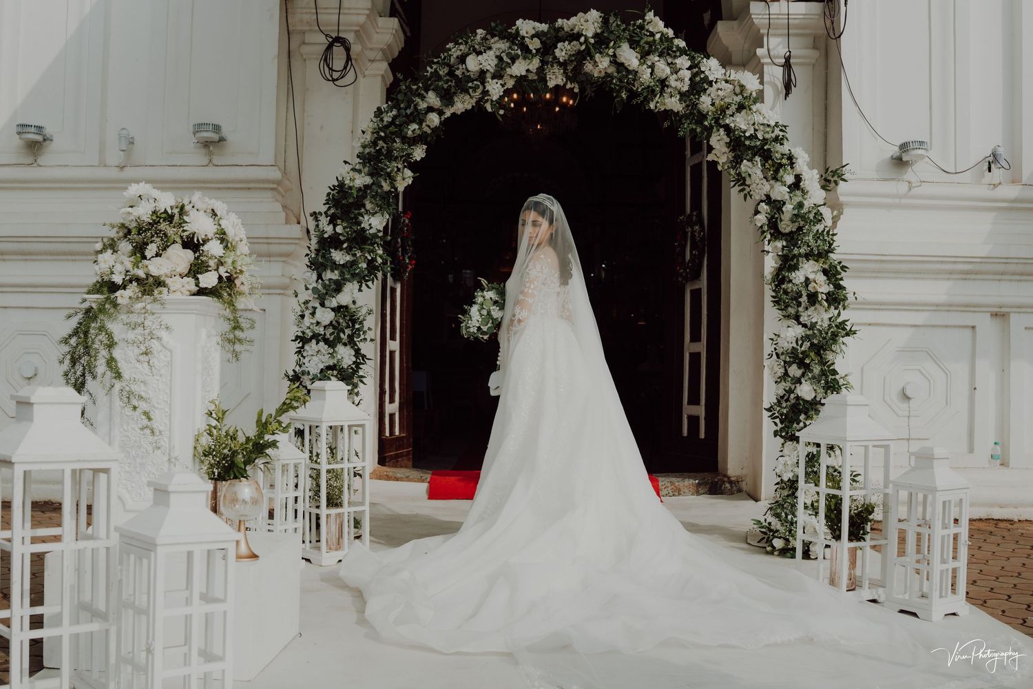 Elegant white wedding ceremony setup with floral arch and flowing cathedral veil in an ornate indoor venue.