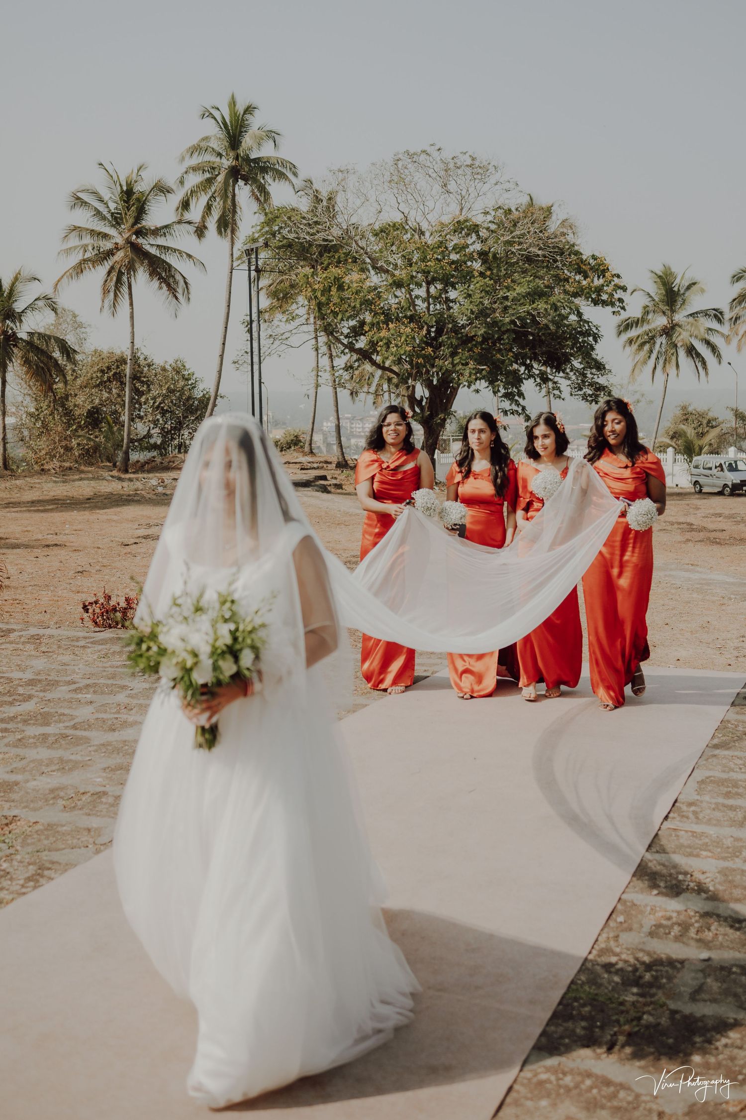 Bride in flowing white gown with bridesmaids in red dresses holding veil on tropical beach with palm trees in background.