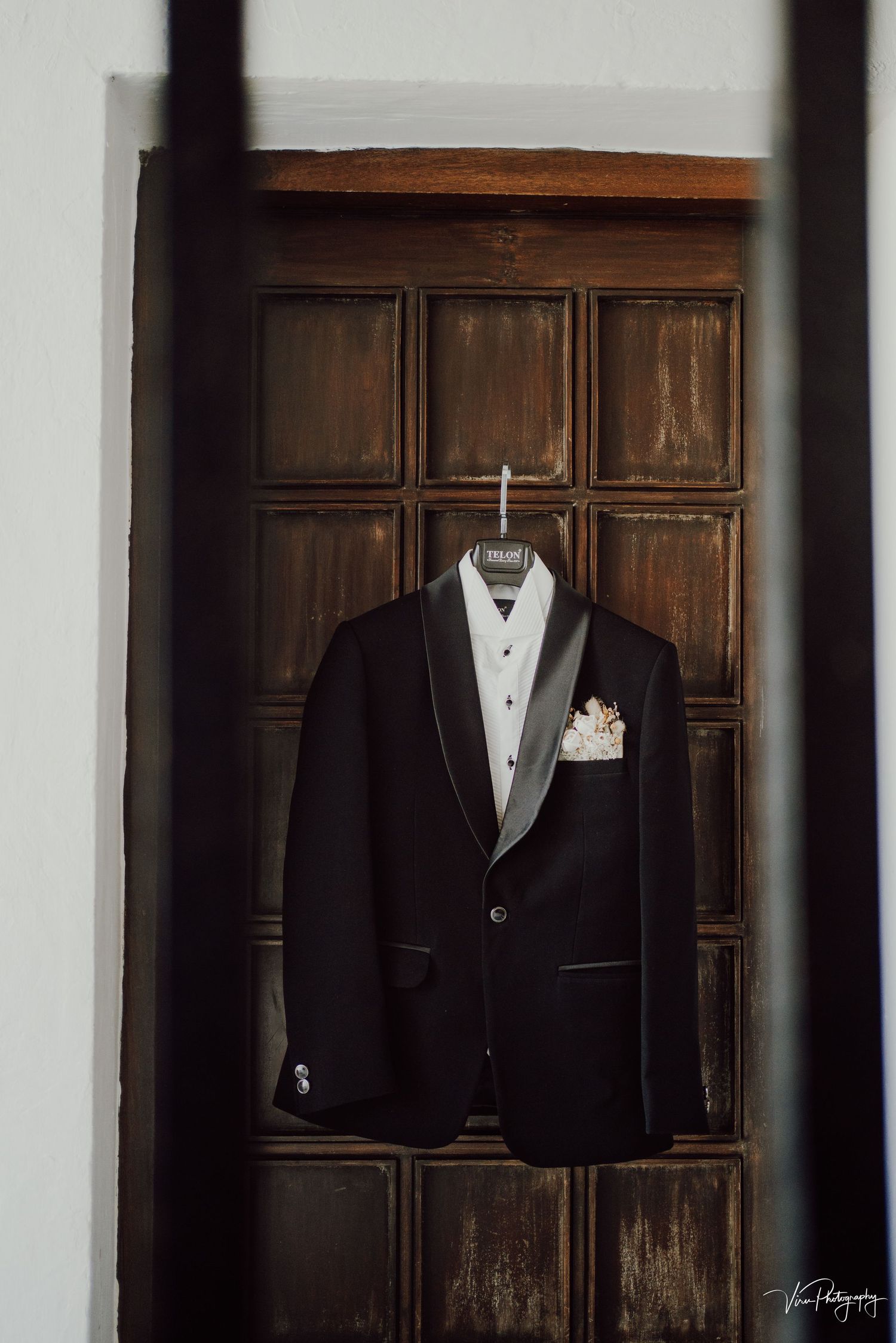 An elegant black tuxedo jacket with satin lapels and a pocket square hangs on display against a dark wooden paneled wall.