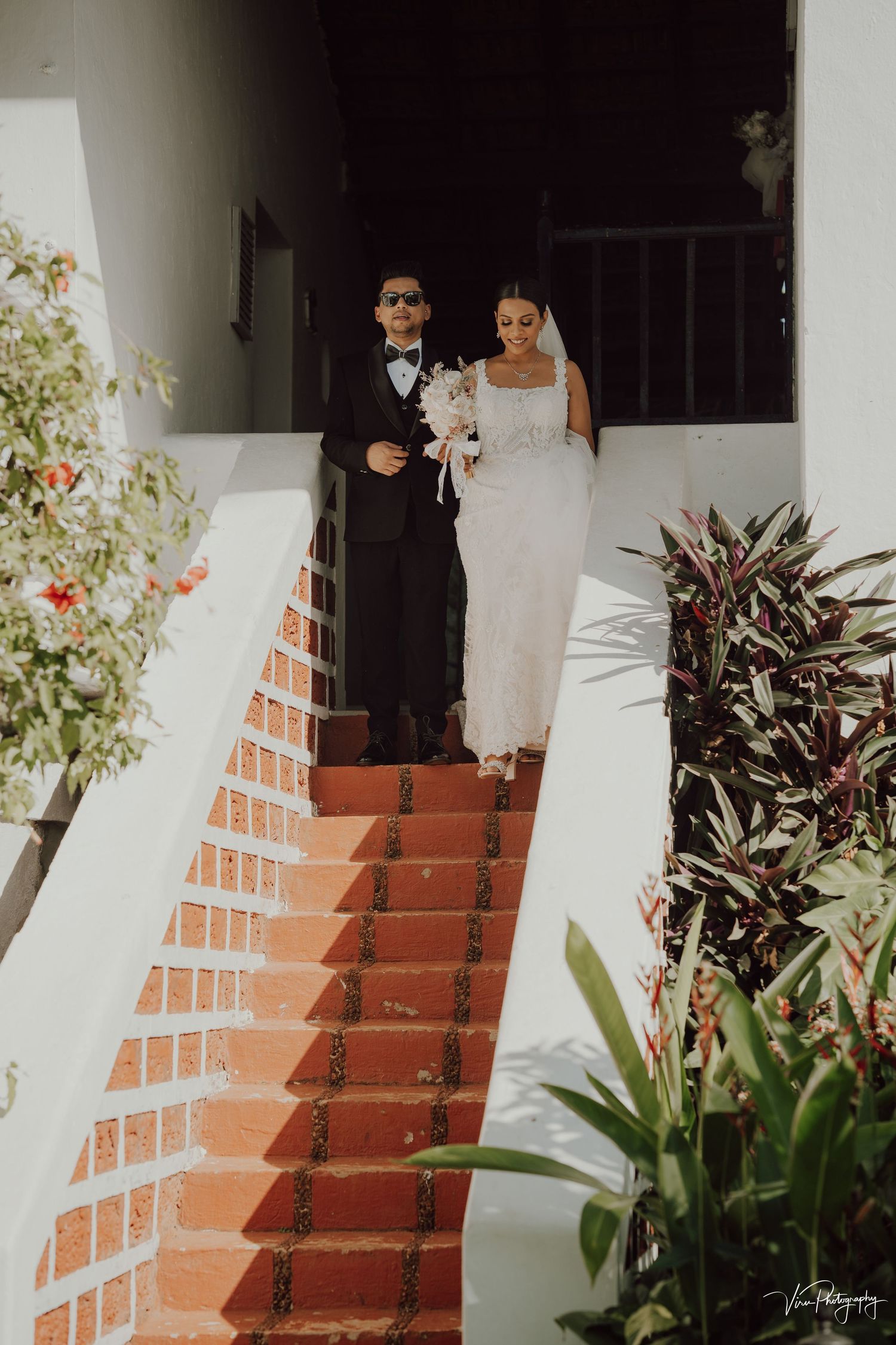 A couple in formal wedding attire descend elegant terracotta stairs lined with lush tropical plants and flowers.