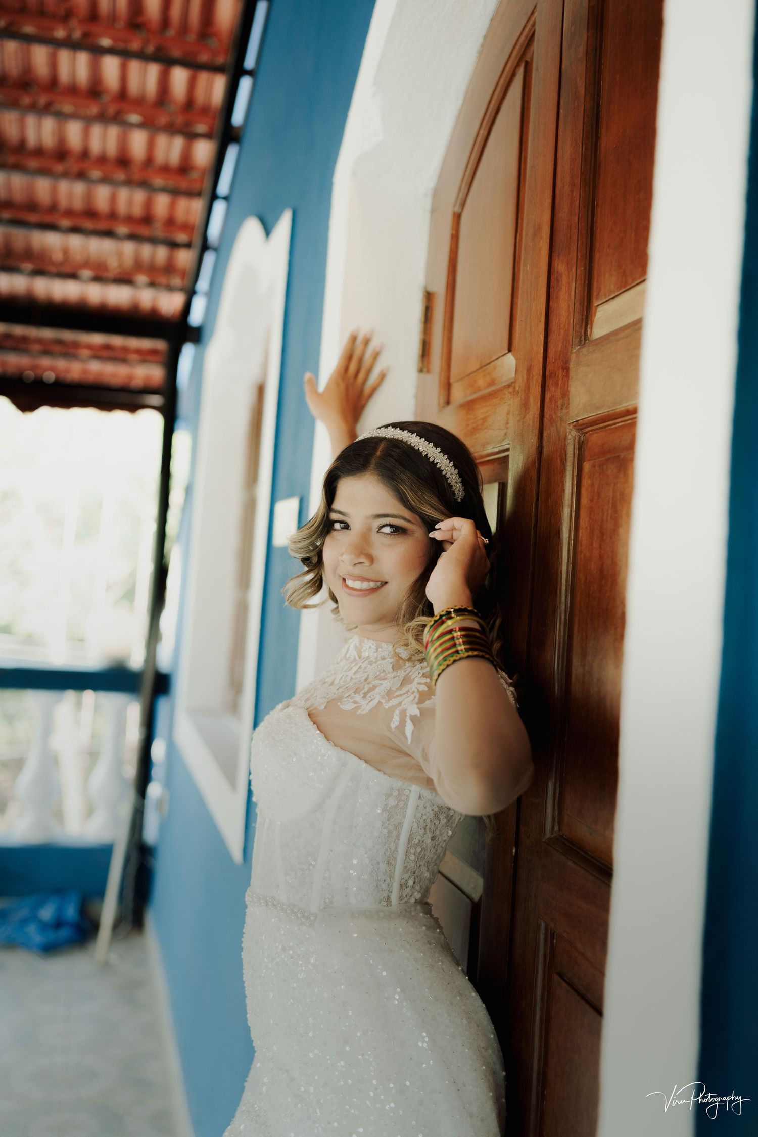 Bride in elegant white lace wedding dress and headpiece poses against blue wall and wooden door.