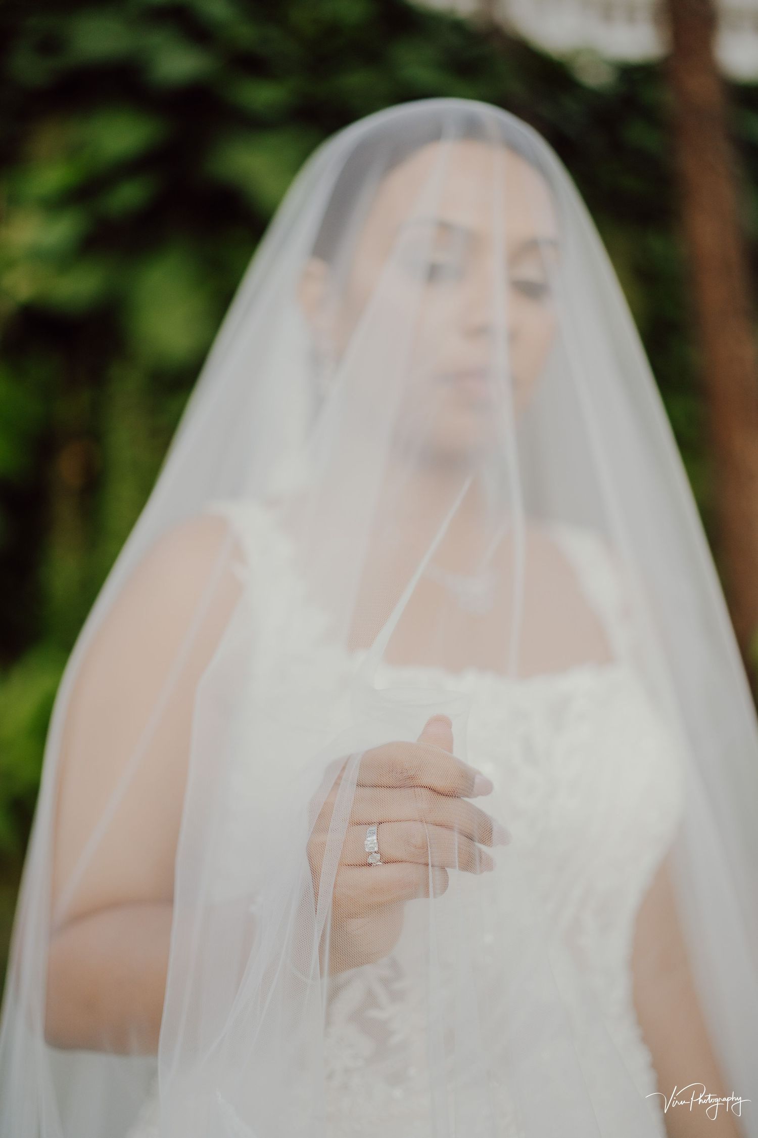 A dreamy bridal portrait through a sheer white veil shows an elegant wedding gown and sparkling engagement ring.