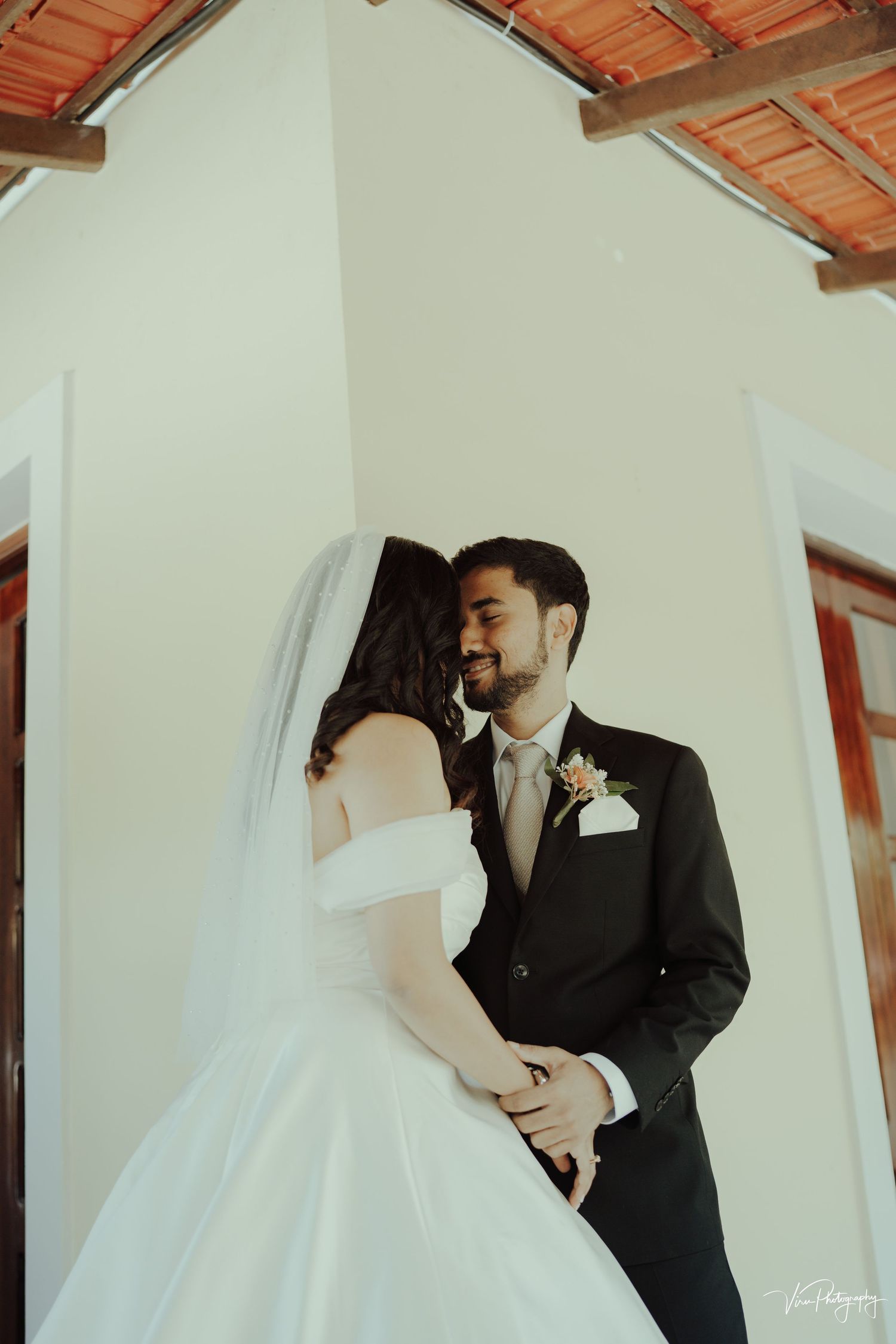 Romantic indoor wedding scene with couple in white dress and black suit standing against light colored wall under wooden beams.