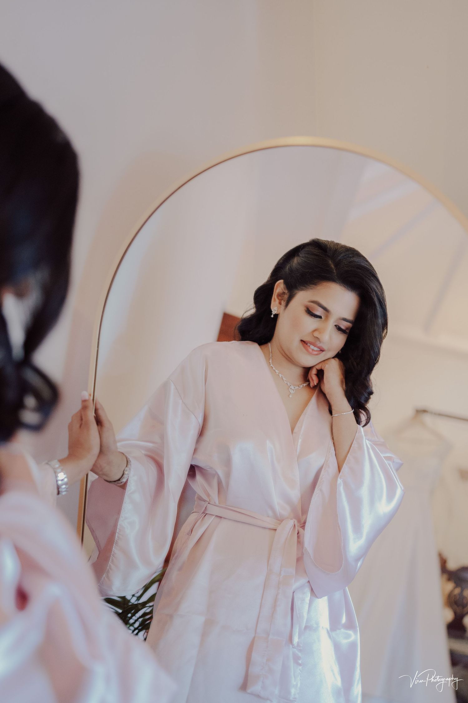 A woman in a pale pink silk robe stands before a mirror with closed eyes and flowing dark hair.