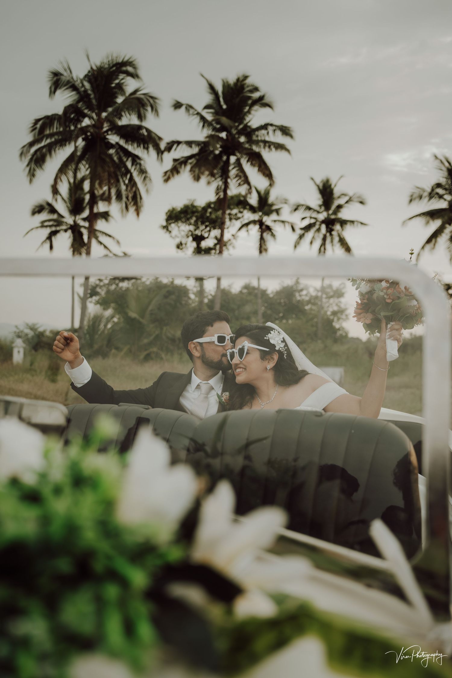 A couple in wedding attire cheering with raised arms while sitting in a car against a tropical palm tree backdrop.