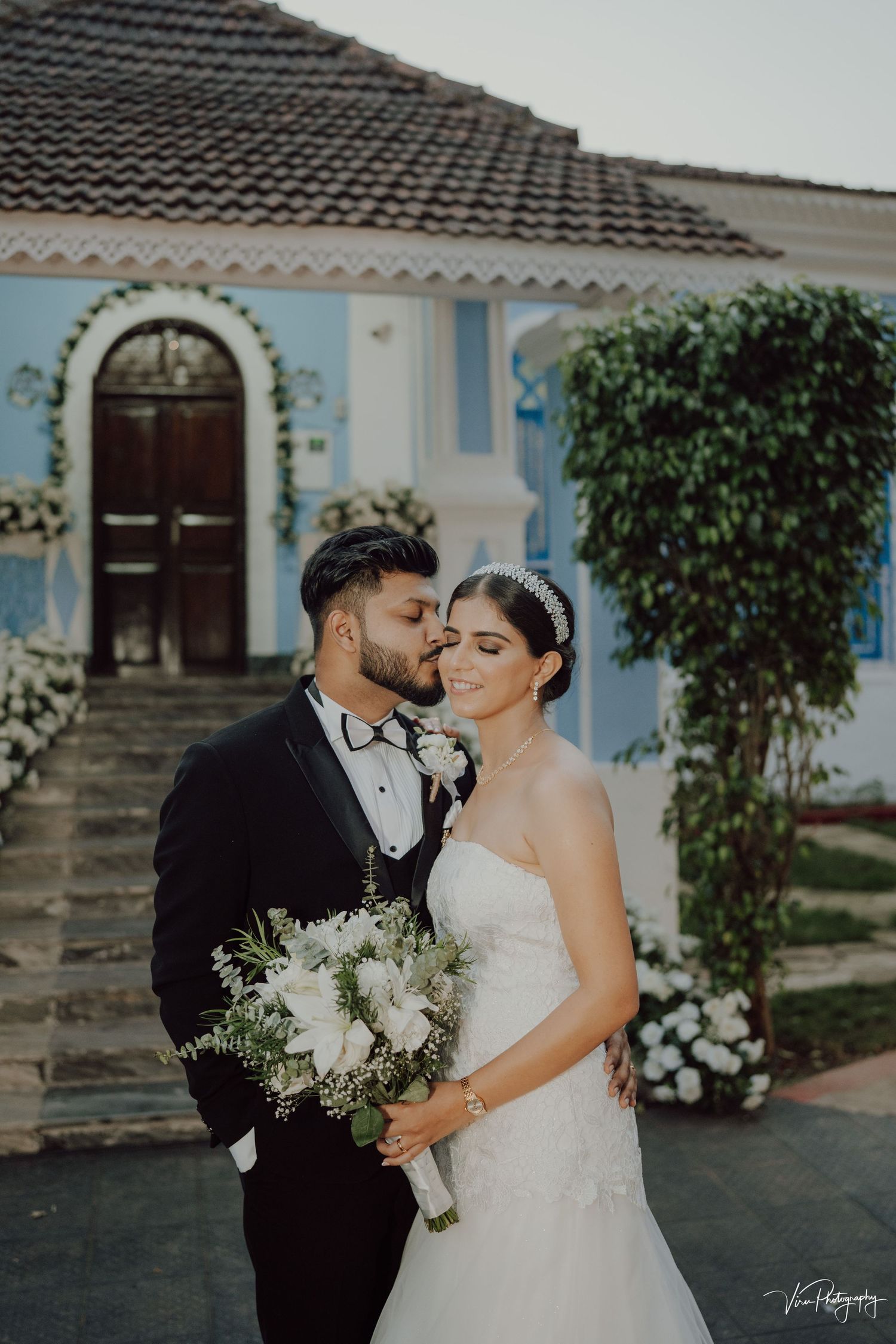 Newlywed couple standing together at blue villa featuring dark wooden door and climbing vines.