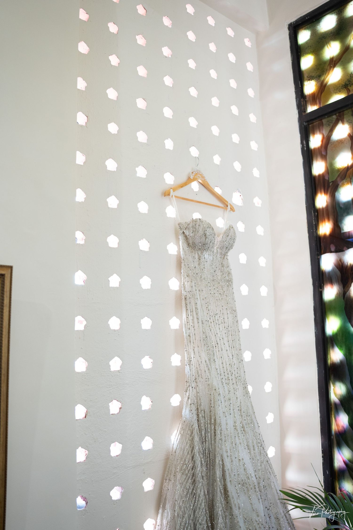 Elegant beaded wedding dress hanging on wooden hanger against white wall with decorative light pattern.