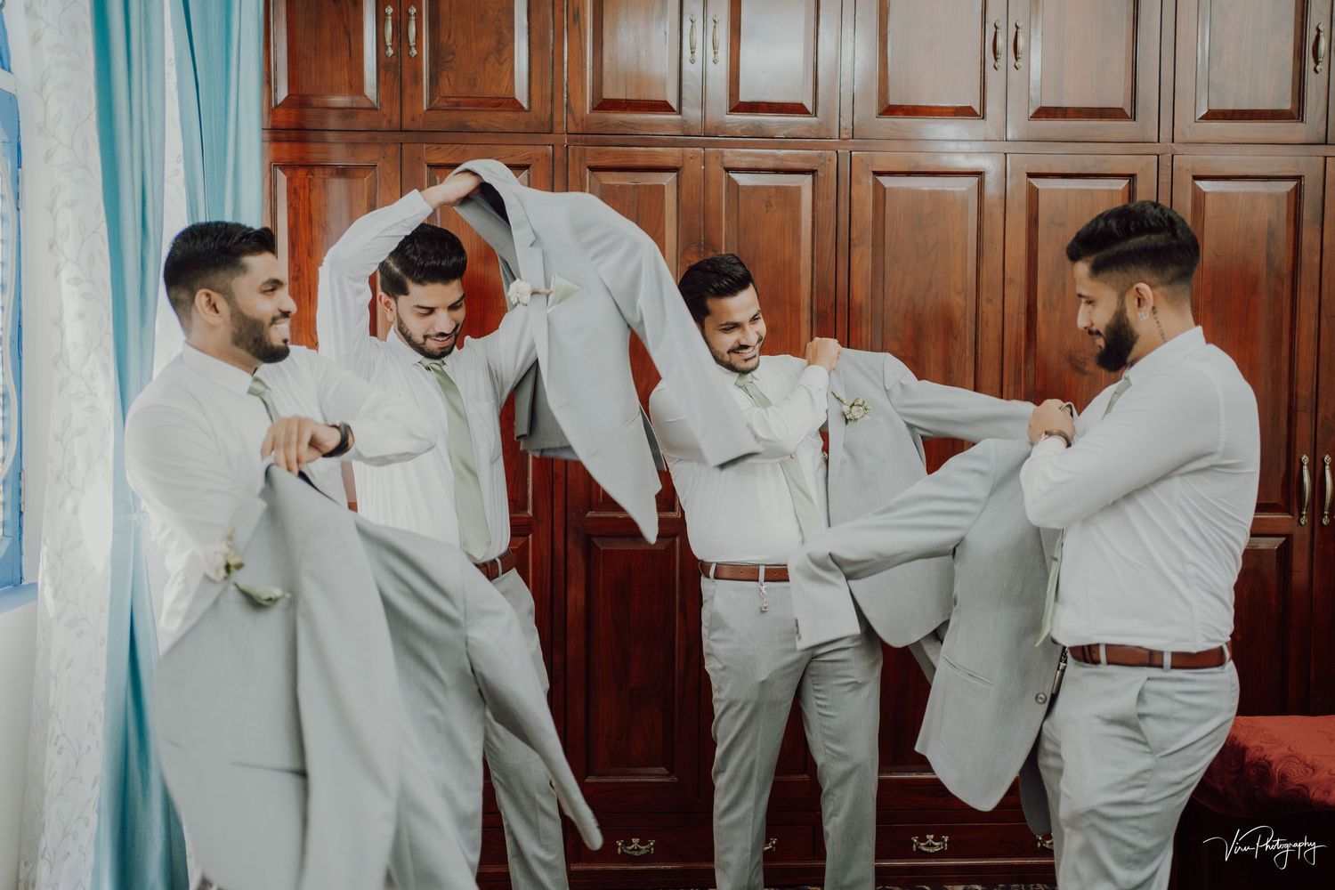Groomsmen in light gray suits mingle and laugh together while adjusting their outfits before a wedding ceremony.