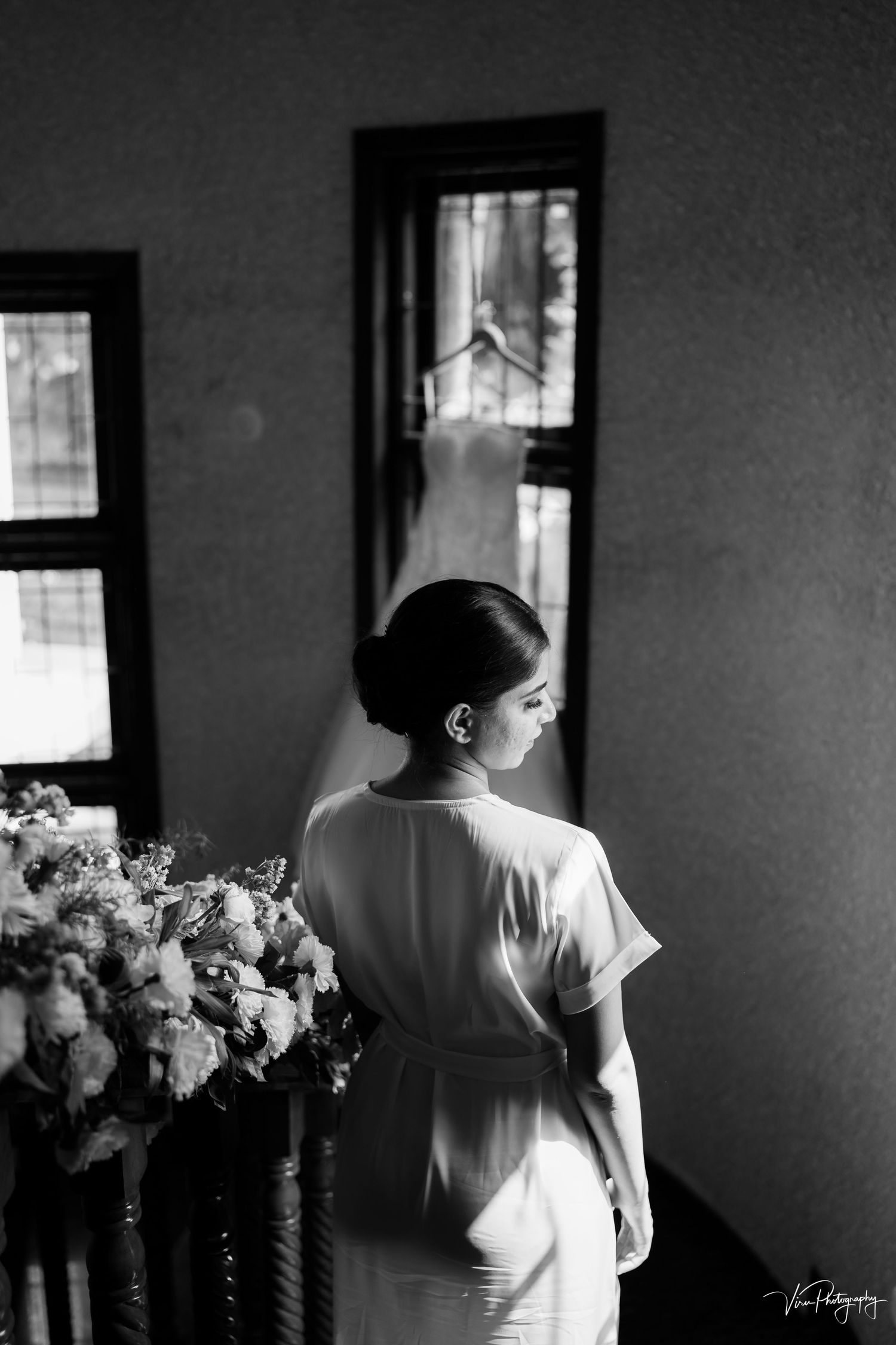 Black and white photograph of a person in a white dress gazing through a window, with a bouquet of flowers nearby.