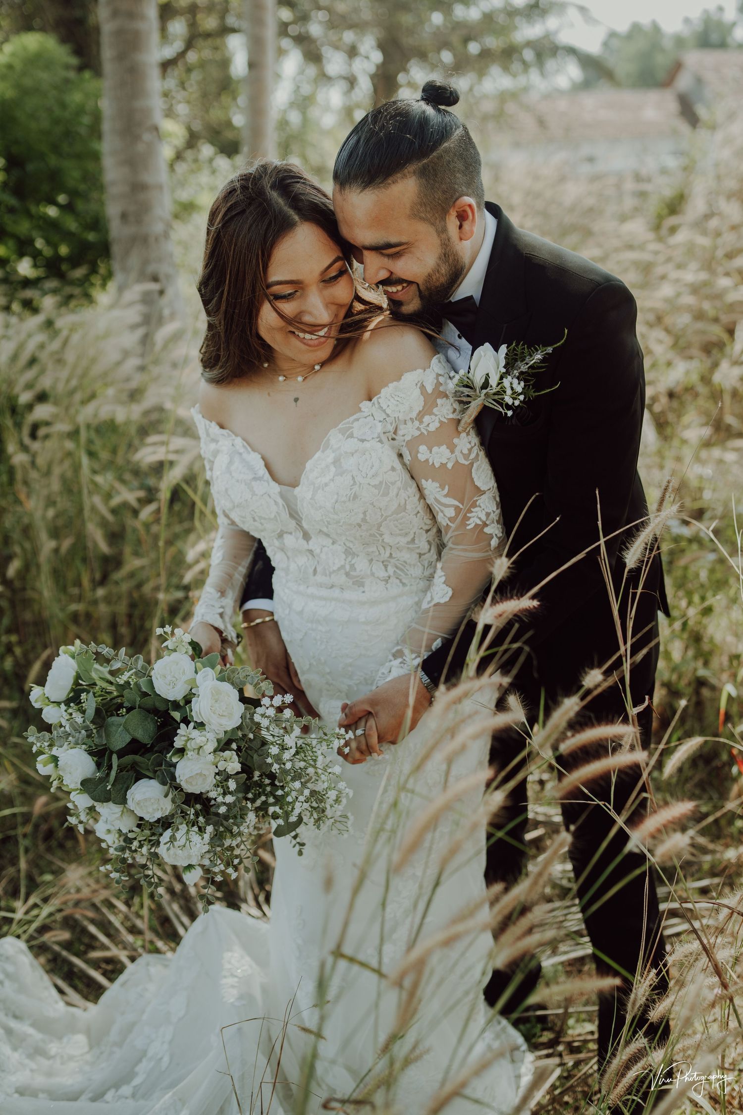 A bride in lace gown and groom in black suit embrace in tall grass during a romantic sunset wedding photoshoot.