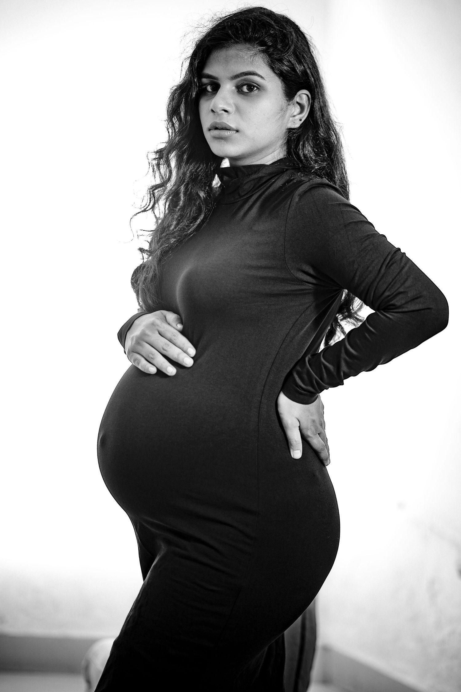 Black and white maternity portrait of pregnant woman in long-sleeved dress posing against light background.