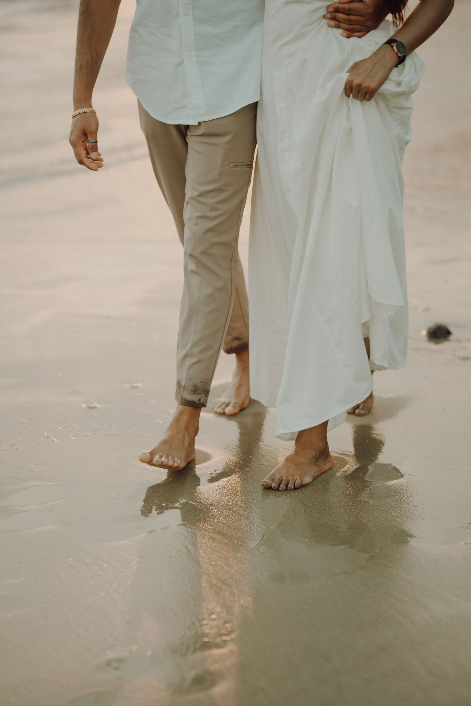Close-up view of barefoot couple standing on wet beach sand in casual beige and white beach attire.
