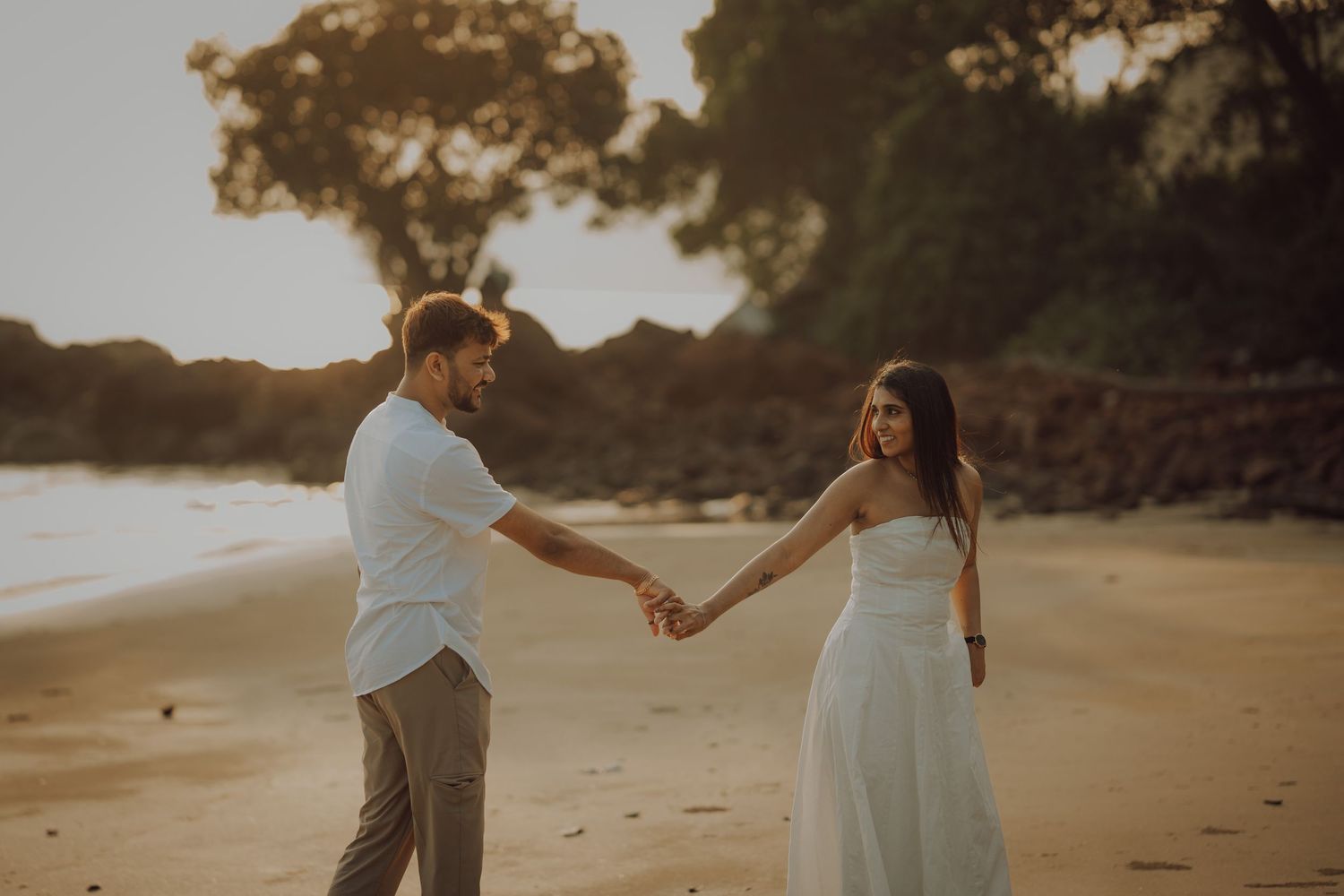 A couple holds hands while walking on a sandy beach at sunset in warm golden light.