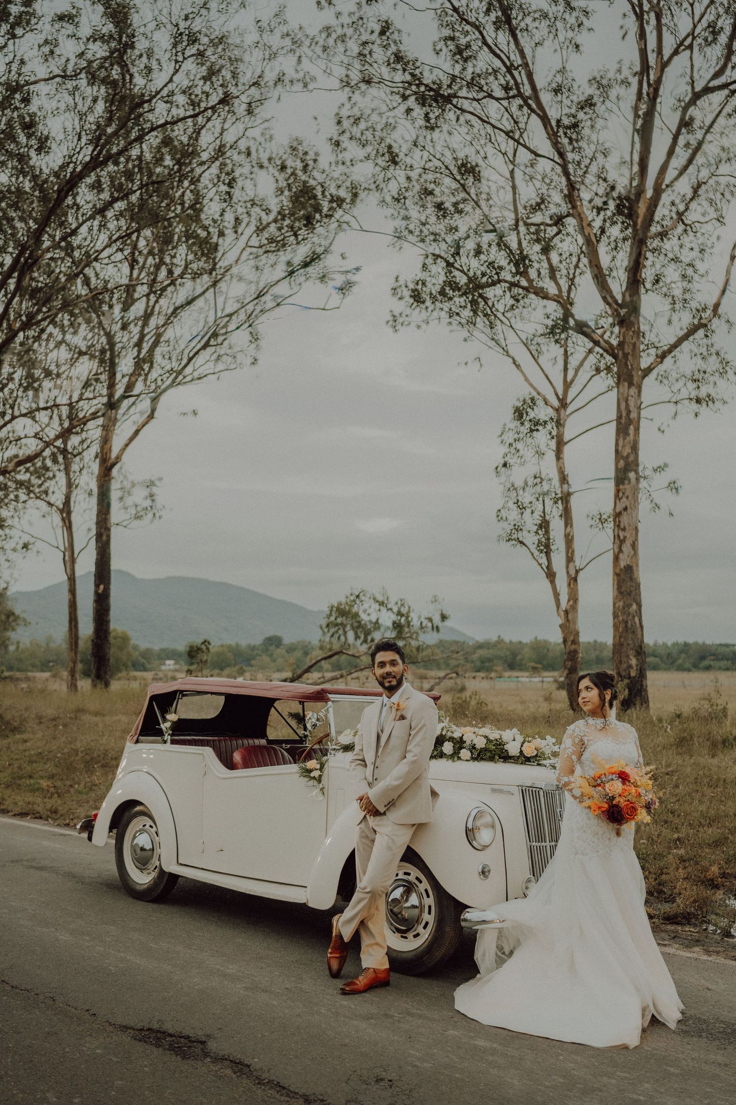 Bride and groom pose beside a vintage white car adorned with flowers on a country road with trees and mountains in the background.