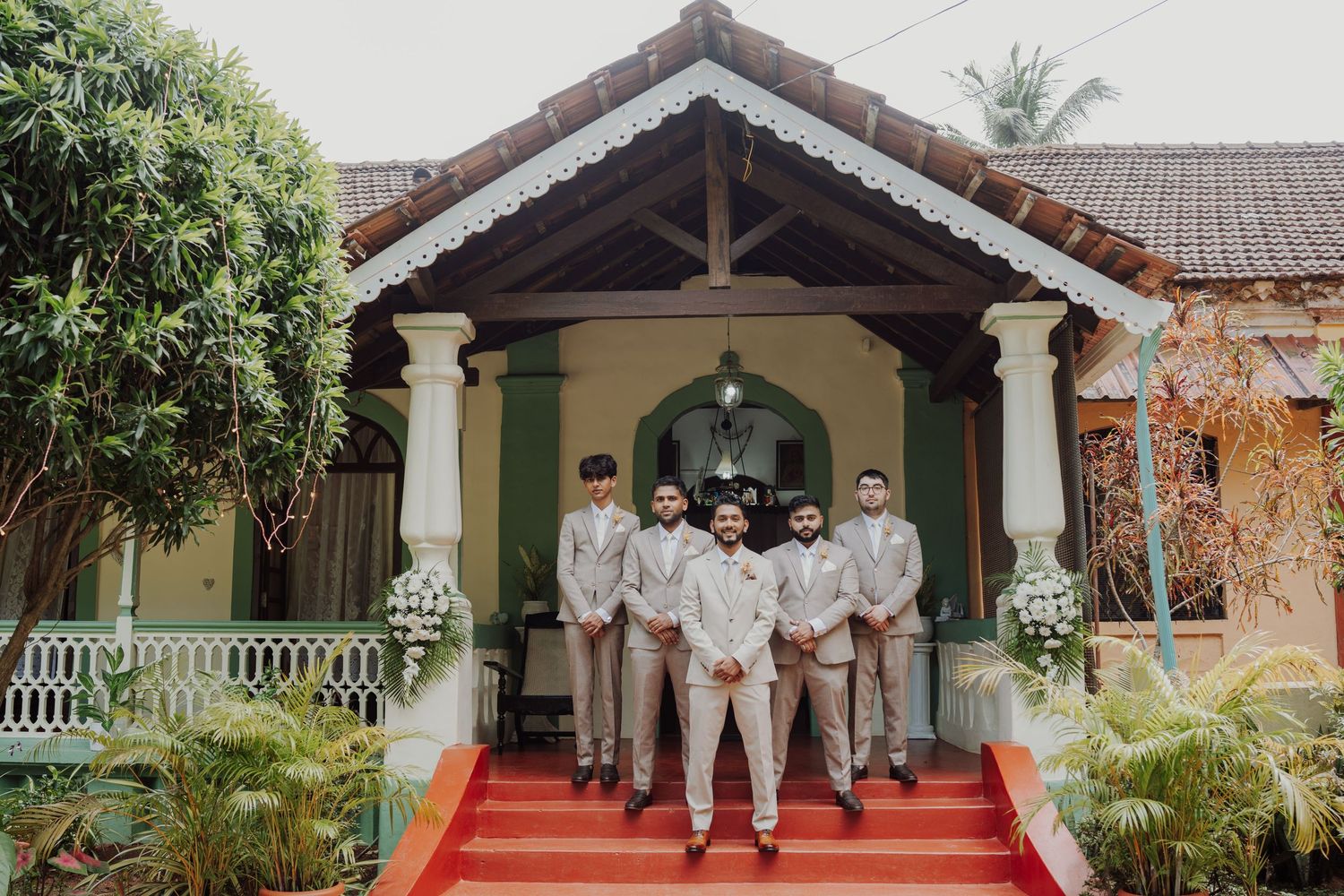 Groomsmen in beige suits pose together on red steps in front of a charming colonial-style building with a tiled roof.