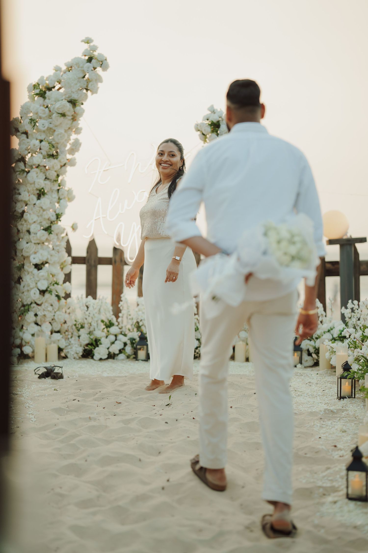 Couple in white attire share a romantic beach proposal moment surrounded by white floral arrangements and candles.