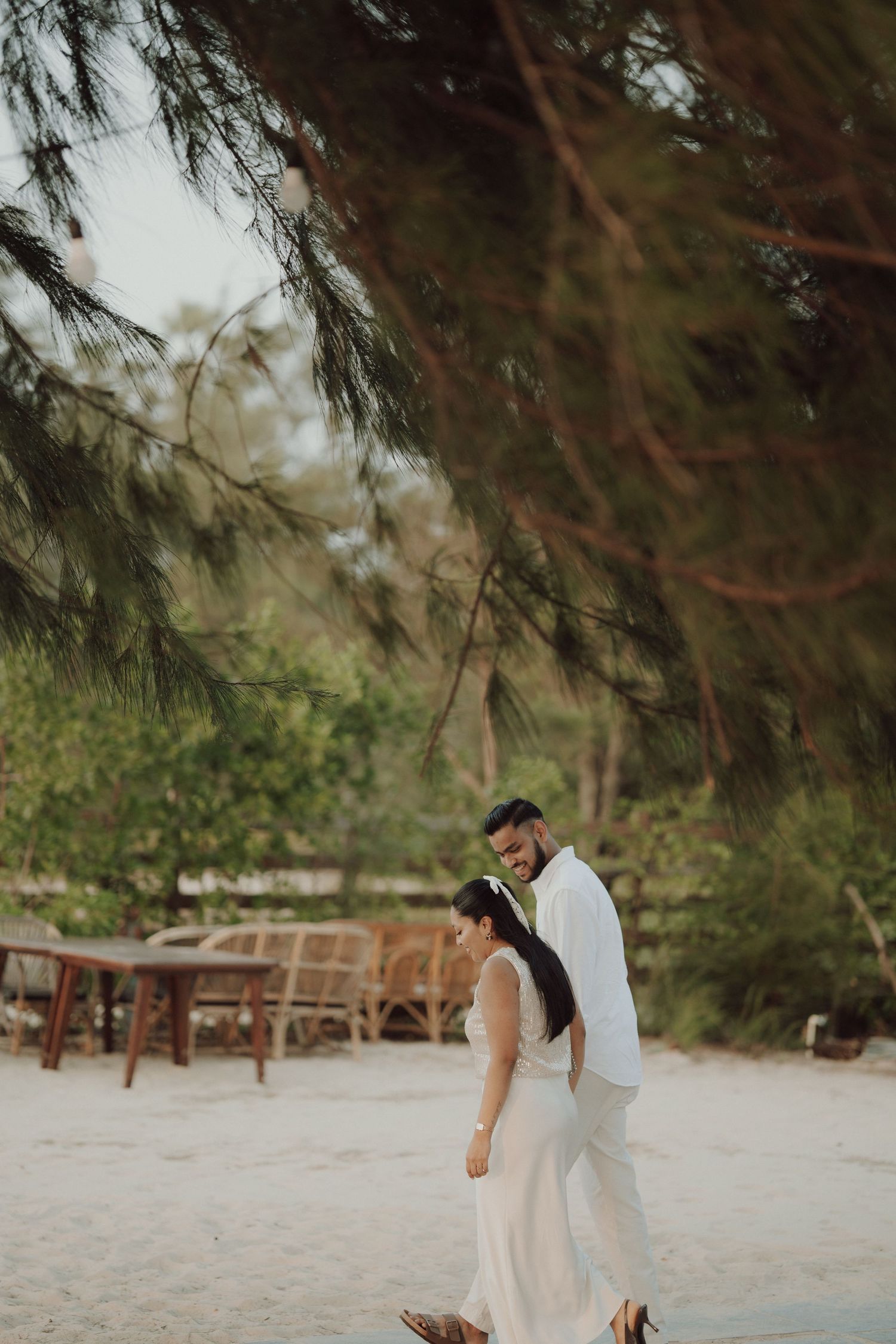 A couple in white wedding attire walks on a sandy beach beneath pine tree branches with lush greenery behind them.