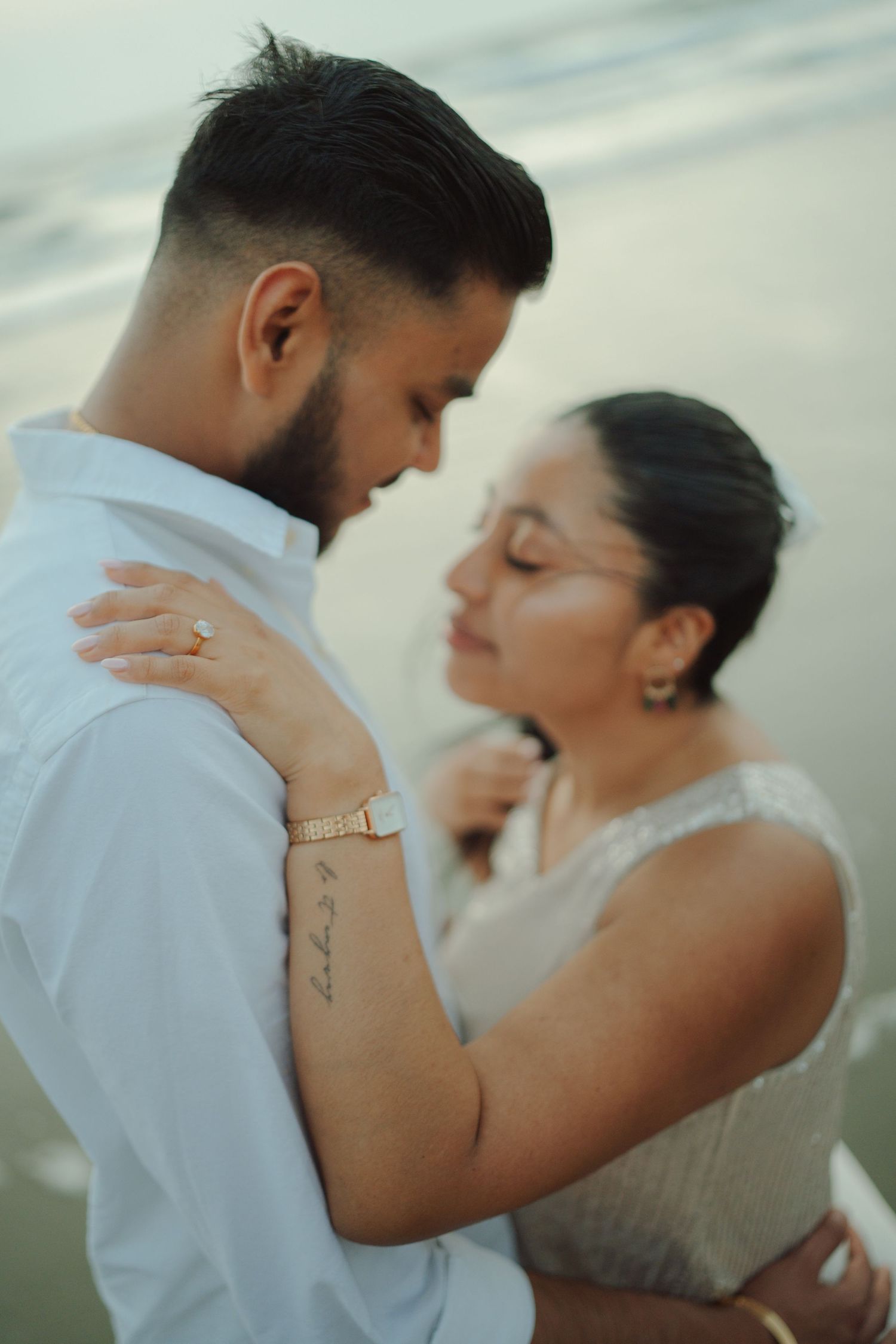 Romantic couple embracing on a beach, woman with tattoo and bracelet, man in white shirt, tender moment.