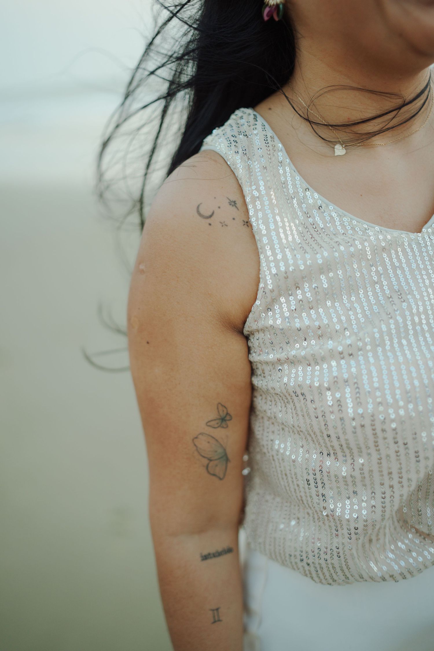 Woman in sequin top with butterfly and moon tattoos on her arm, wearing a delicate necklace.