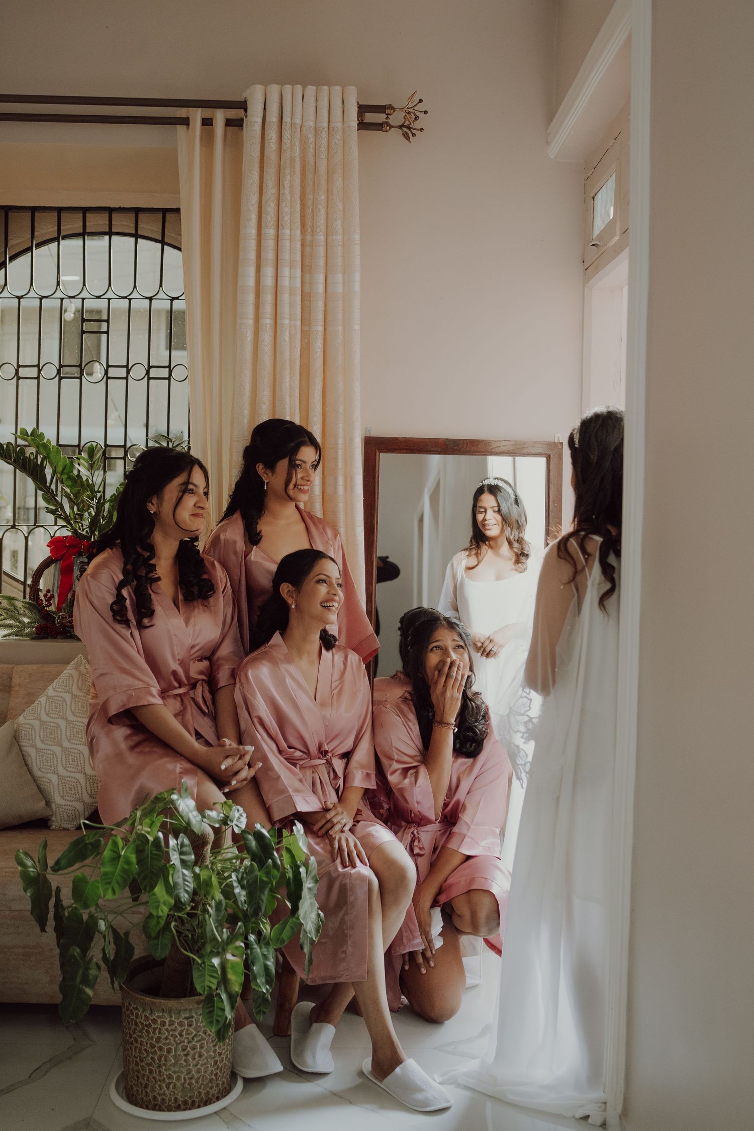 Pink-robed bridesmaids gather in bedroom while bride in white appears in doorway.
