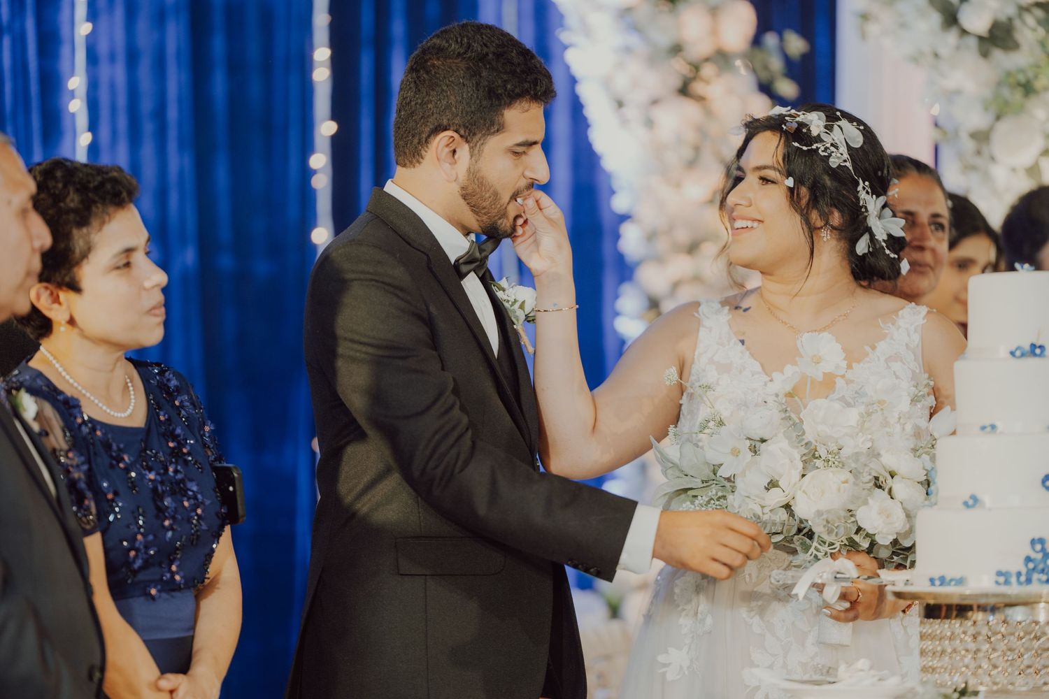 Wedding reception scene with guests gathered around a multi-tiered white cake.