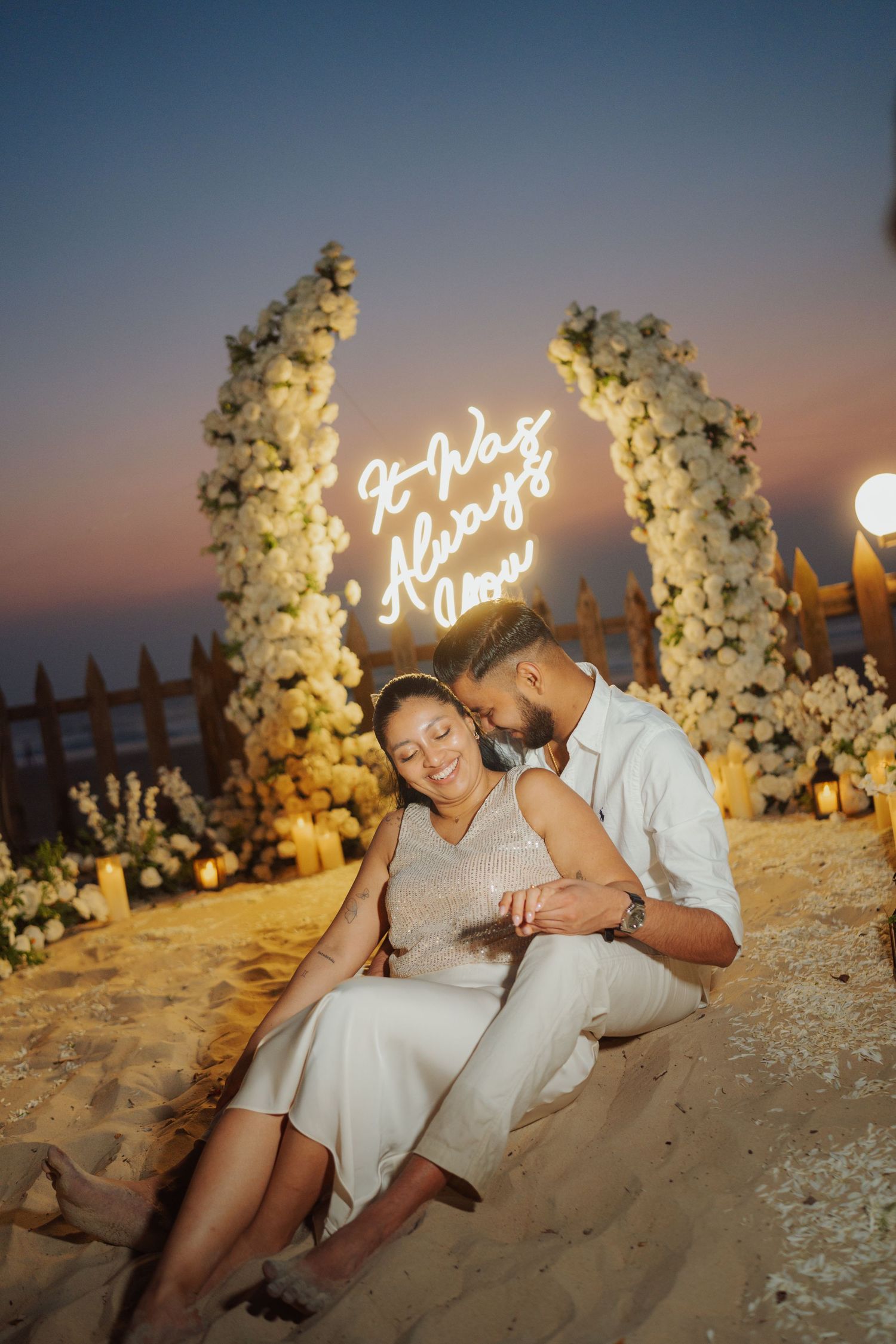 Romantic couple sitting on beach sand at sunset near floral arch with neon sign reading It Was Always You.