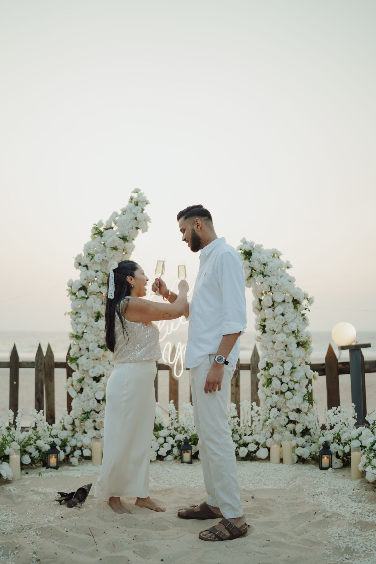 Couple in white attire toasting champagne by a floral arch on a beach at sunset.