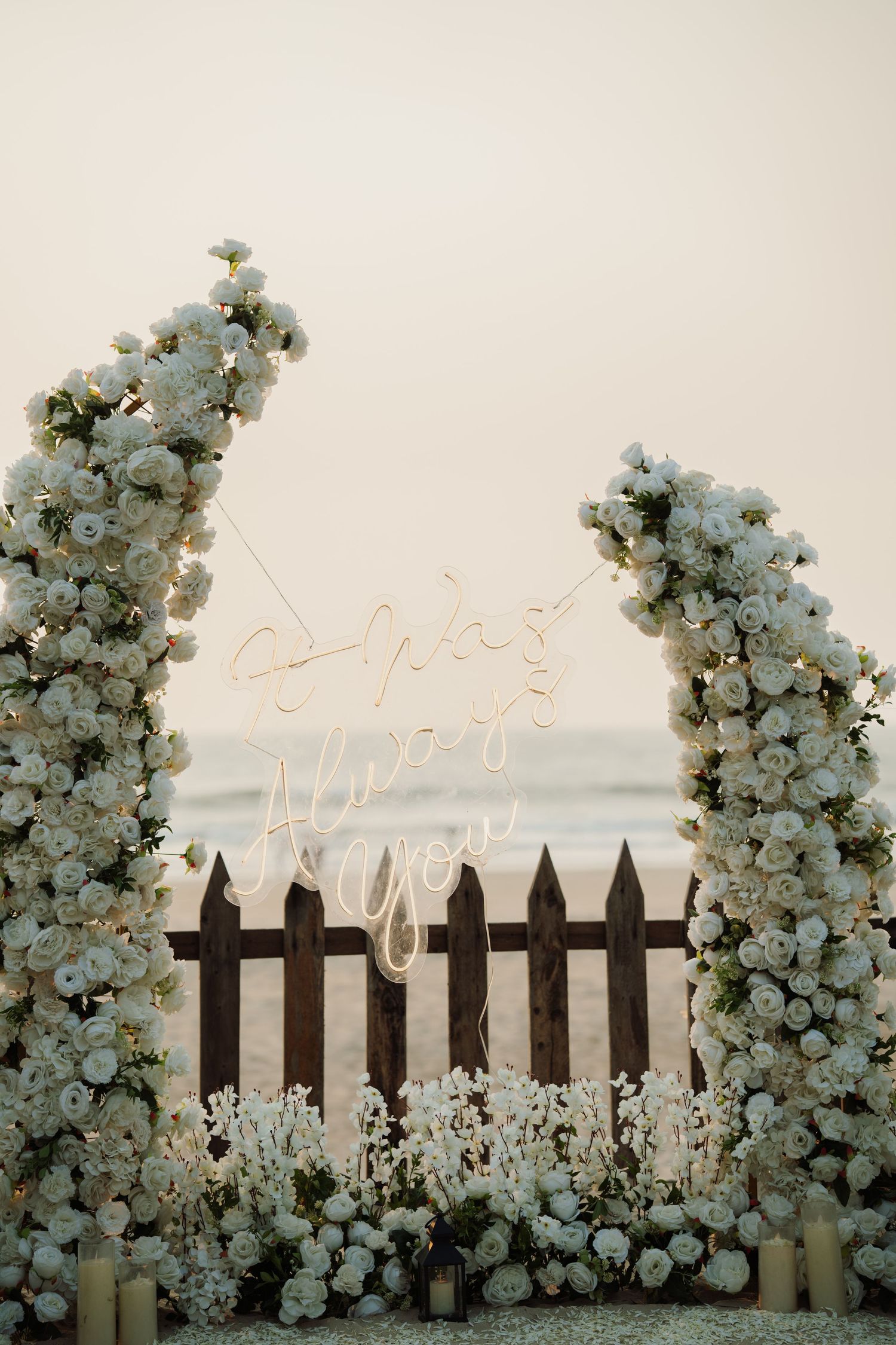 Elegant beach wedding arch adorned with white roses and neon sign, framed by a rustic wooden picket fence at sunset.