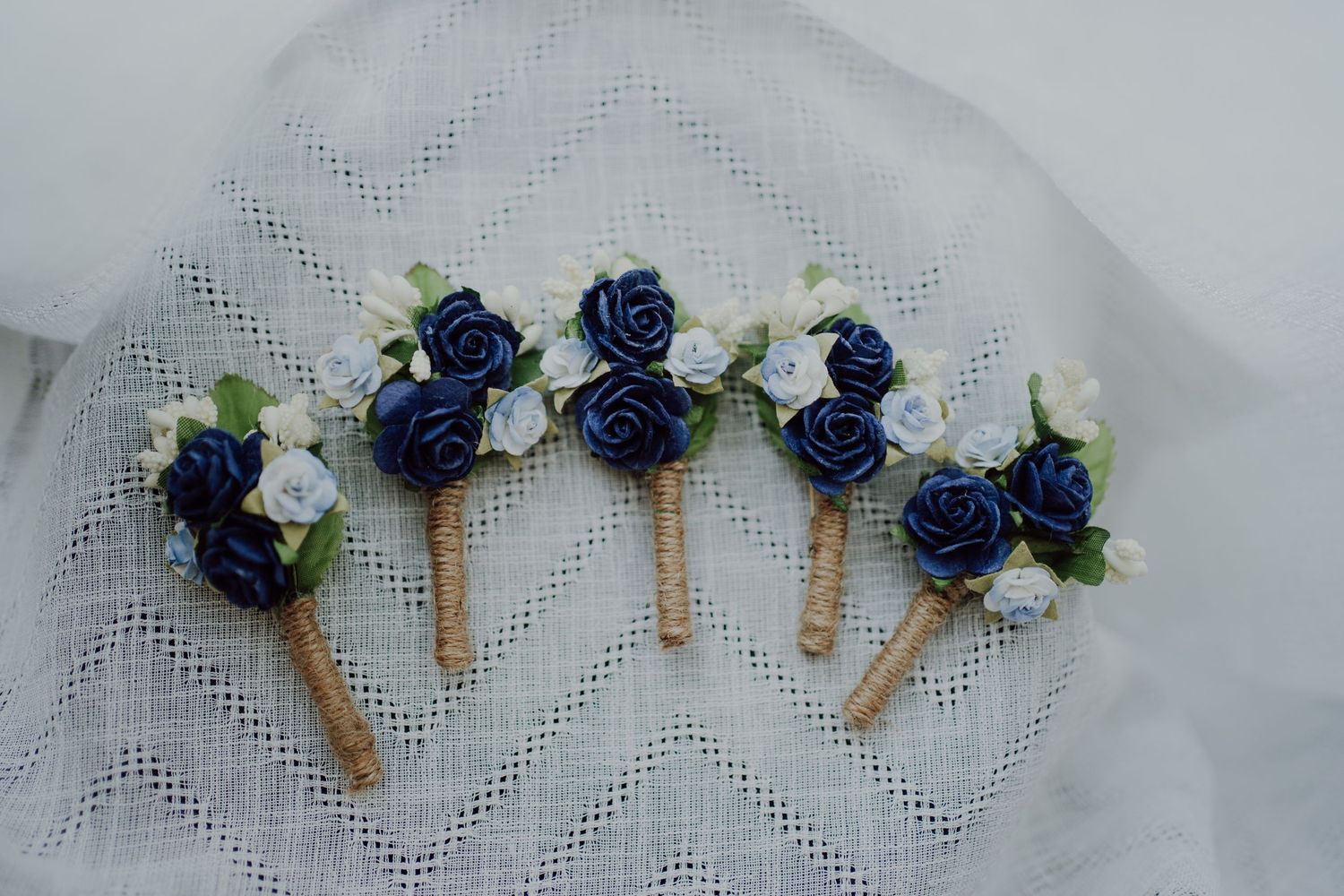 Four elegant boutonnieres with navy blue and white roses arranged on delicate lace fabric.