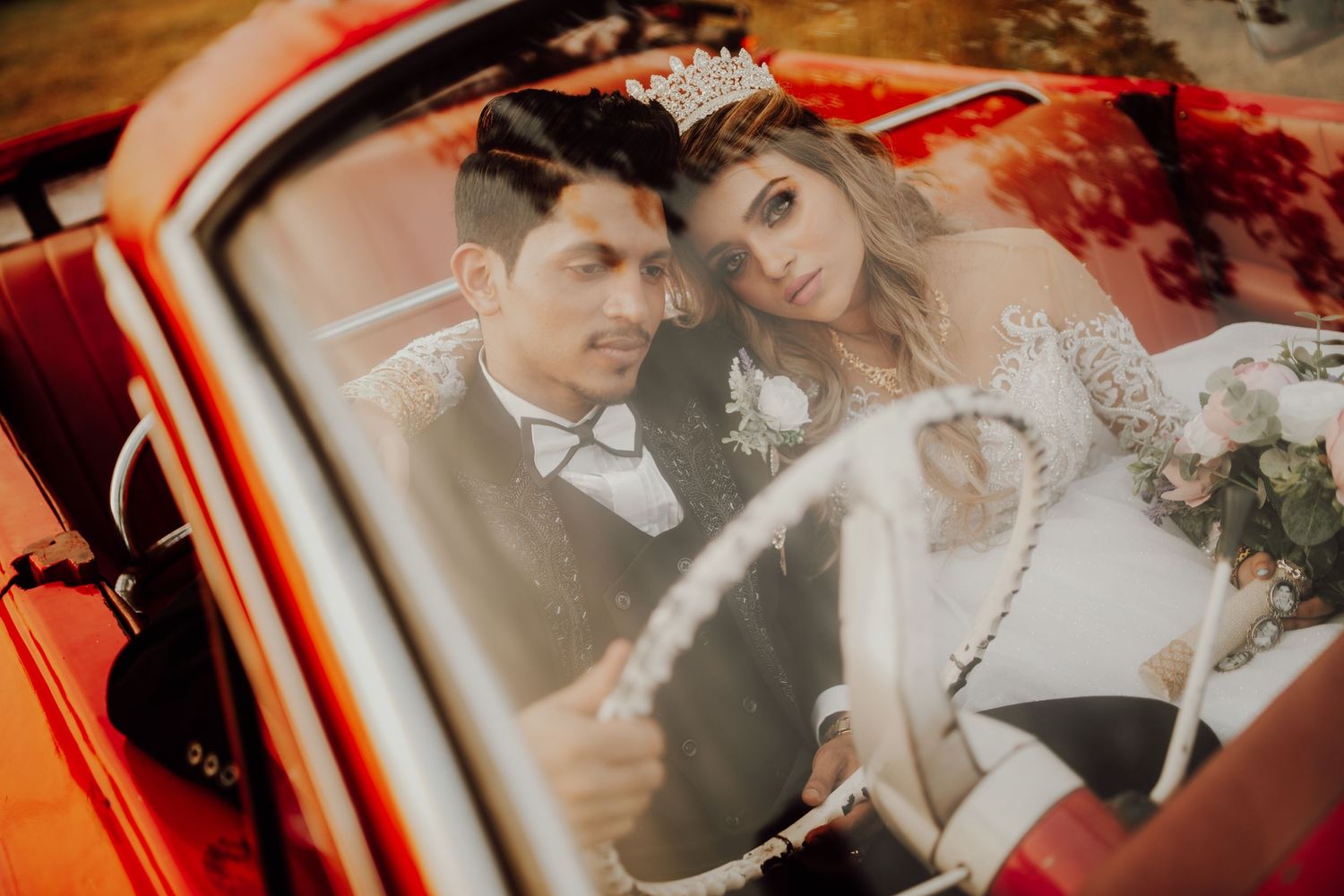 A newly married couple poses together in a vintage red car with romantic autumn lighting and wedding attire.