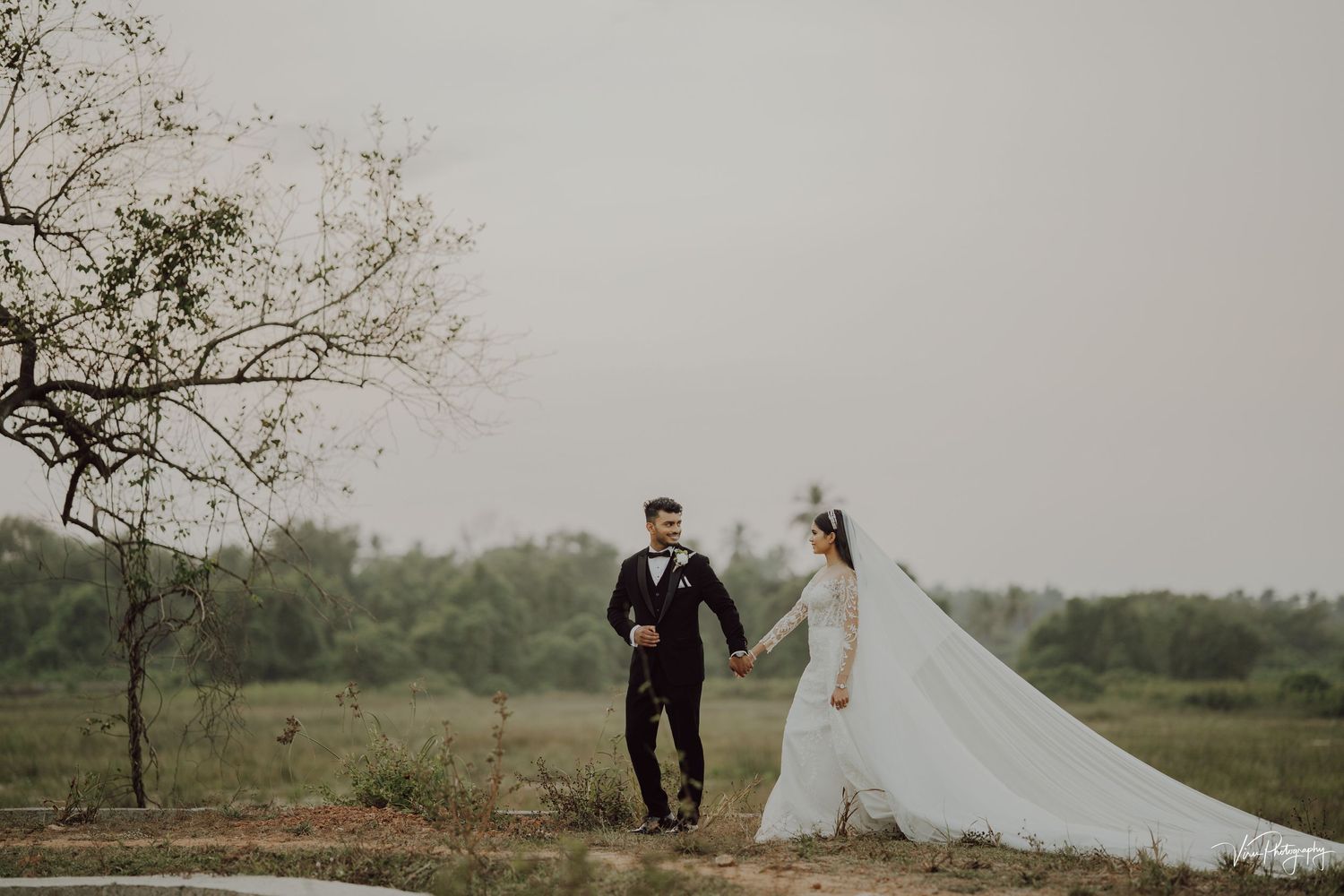 A couple in wedding attire holds hands while walking together in a misty outdoor setting with trees and greenery.