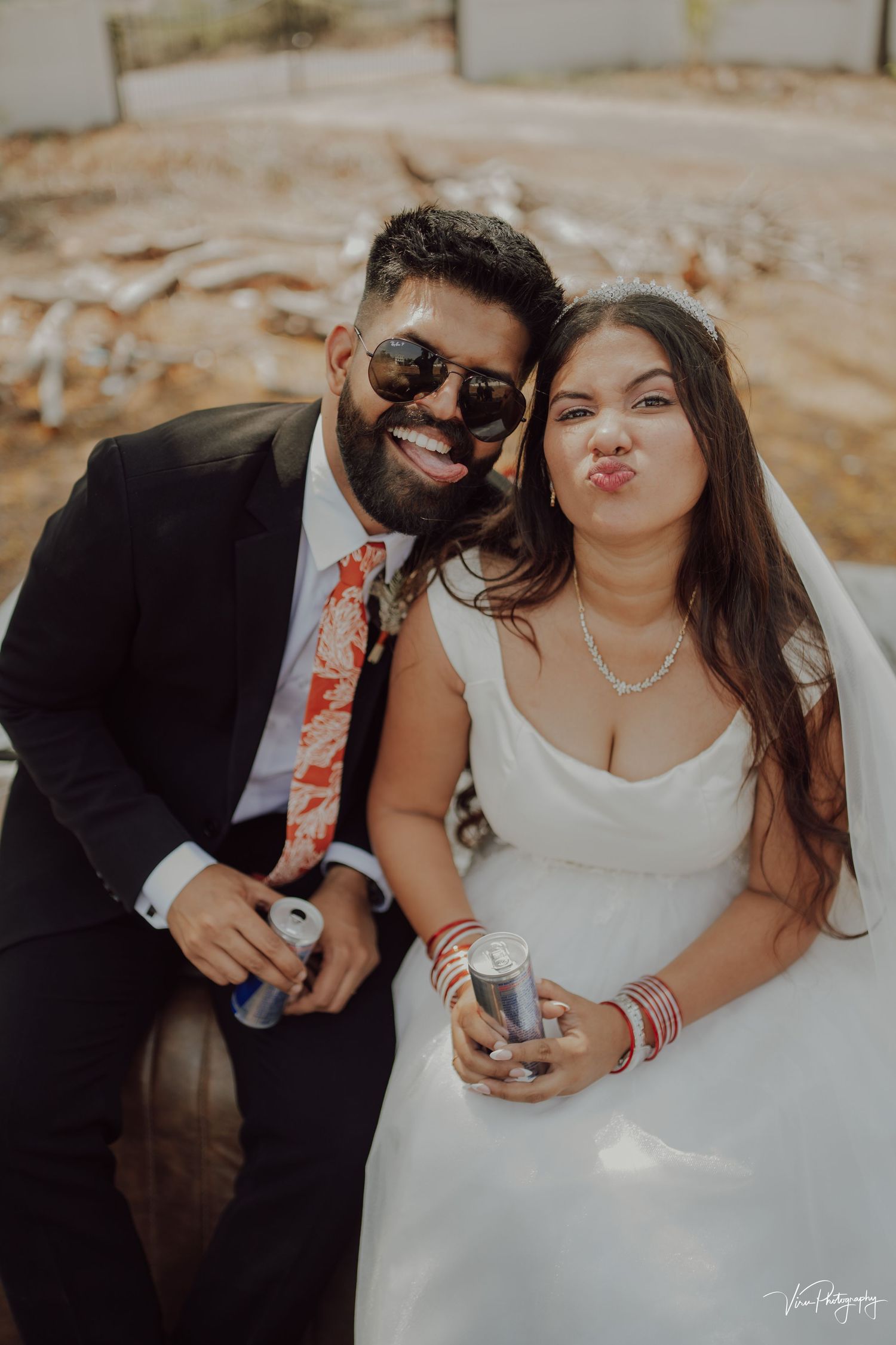 Stylish newlywed couple making silly faces while holding drinks at their outdoor wedding celebration.