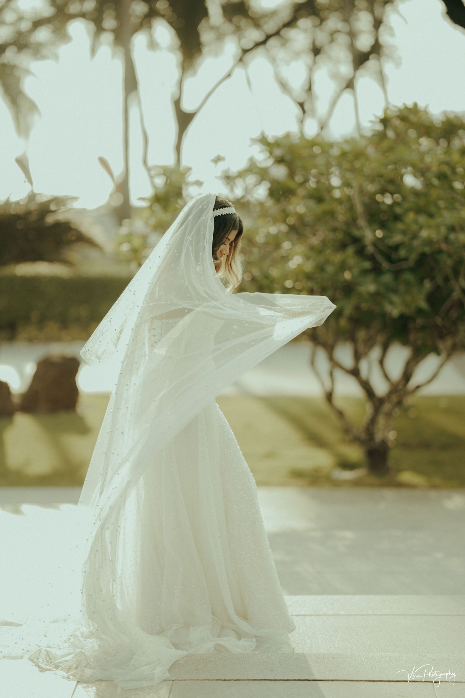 A bride in a flowing white wedding dress and veil twirls on a patio surrounded by soft bokeh trees and greenery.