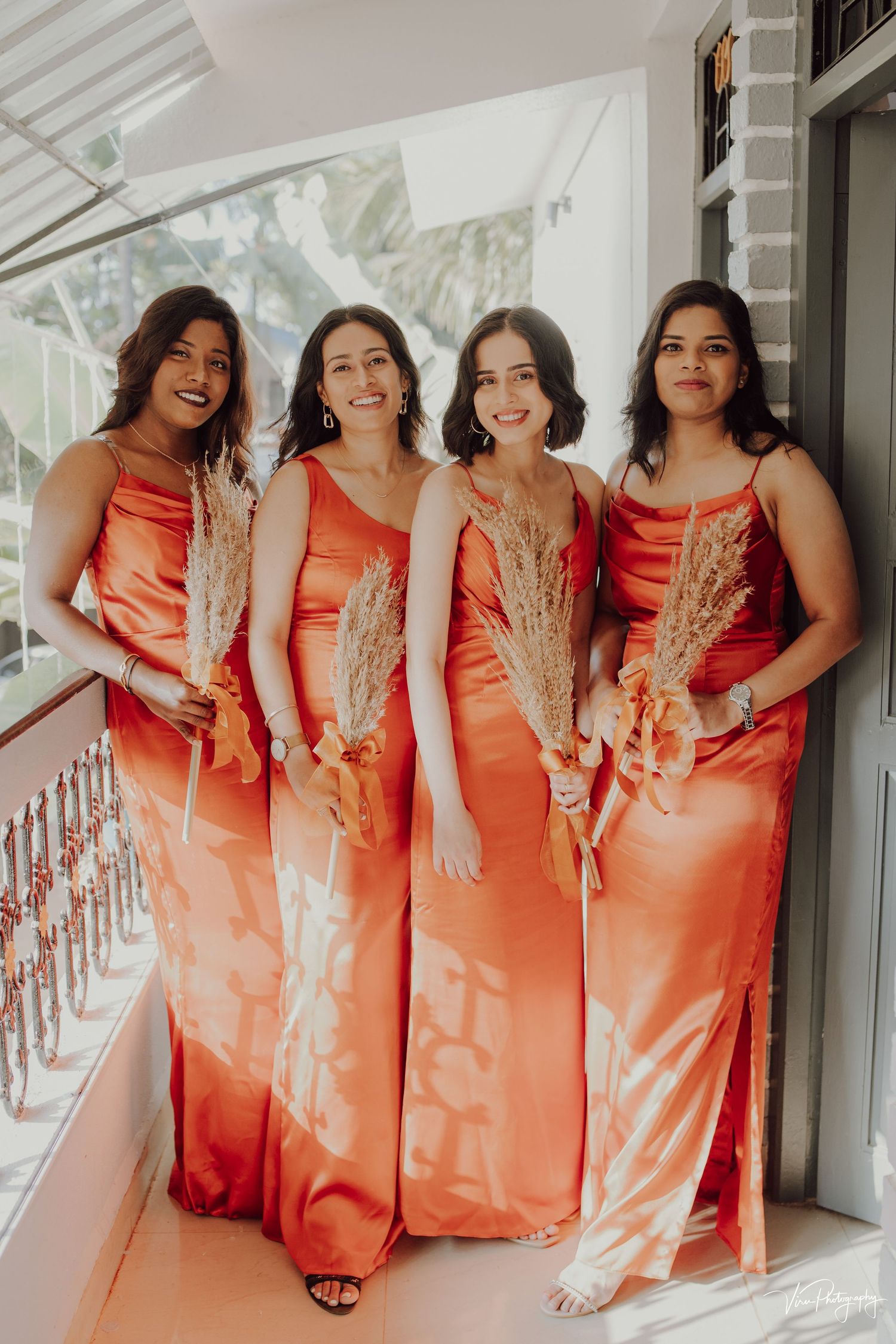 Four women in matching coral satin dresses holding dried palm fronds pose together on a white porch balcony.