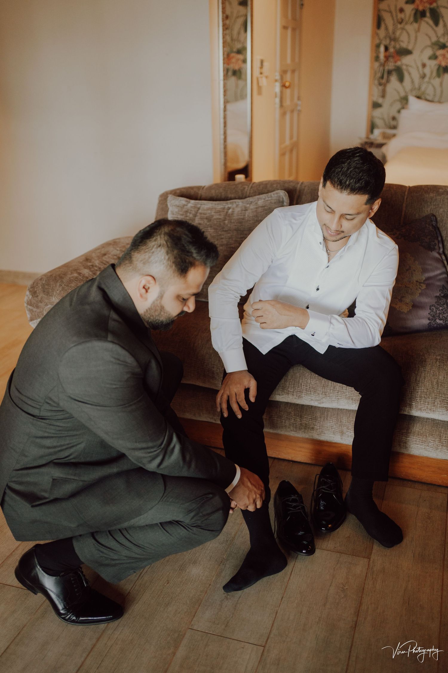 Two people interacting in a room with leather seating, one crouched down adjusting dress shoes while the other sits on a couch.