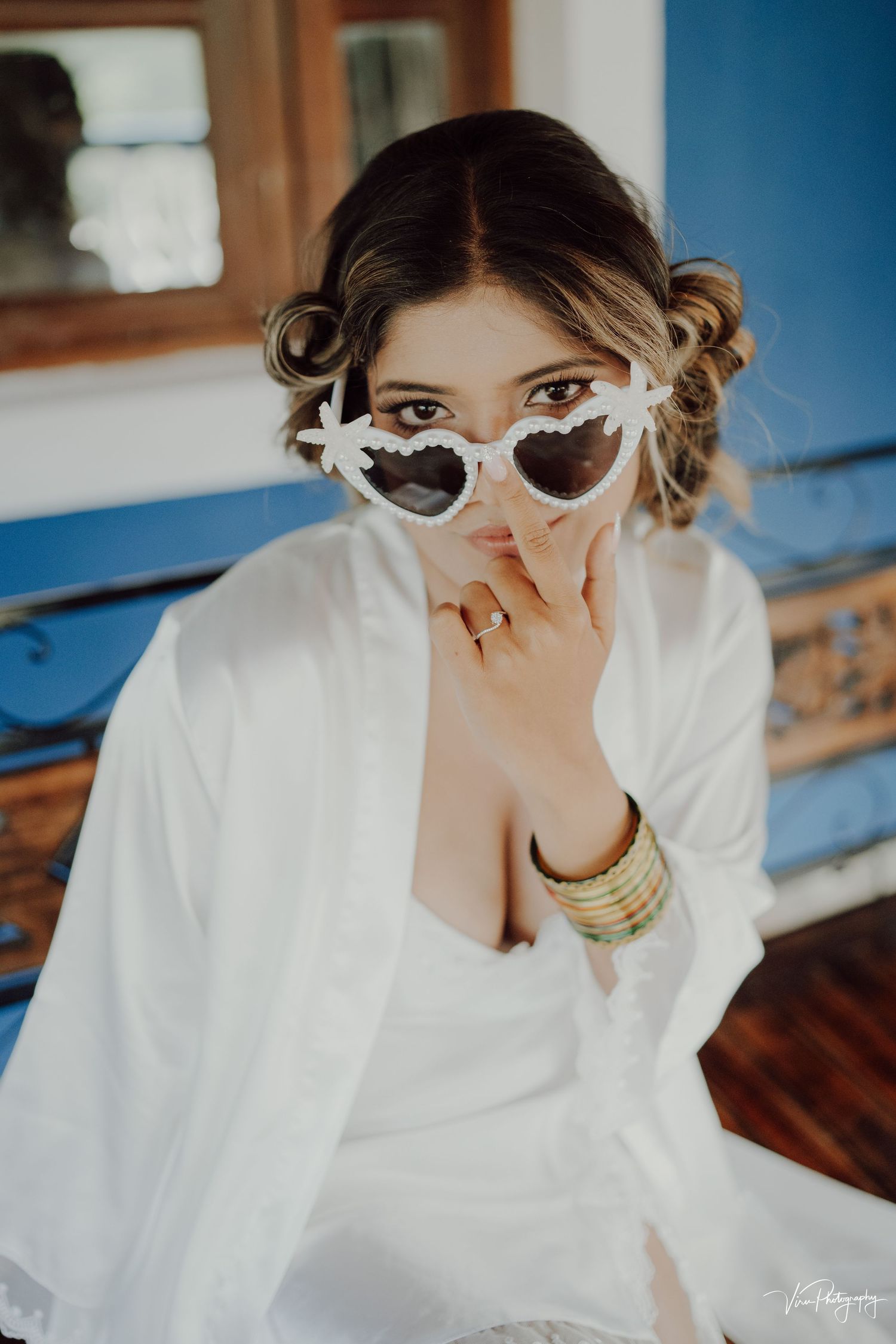 Glamorous bridal portrait featuring heart-shaped sunglasses, white robe and gold bangles in a natural light setting.