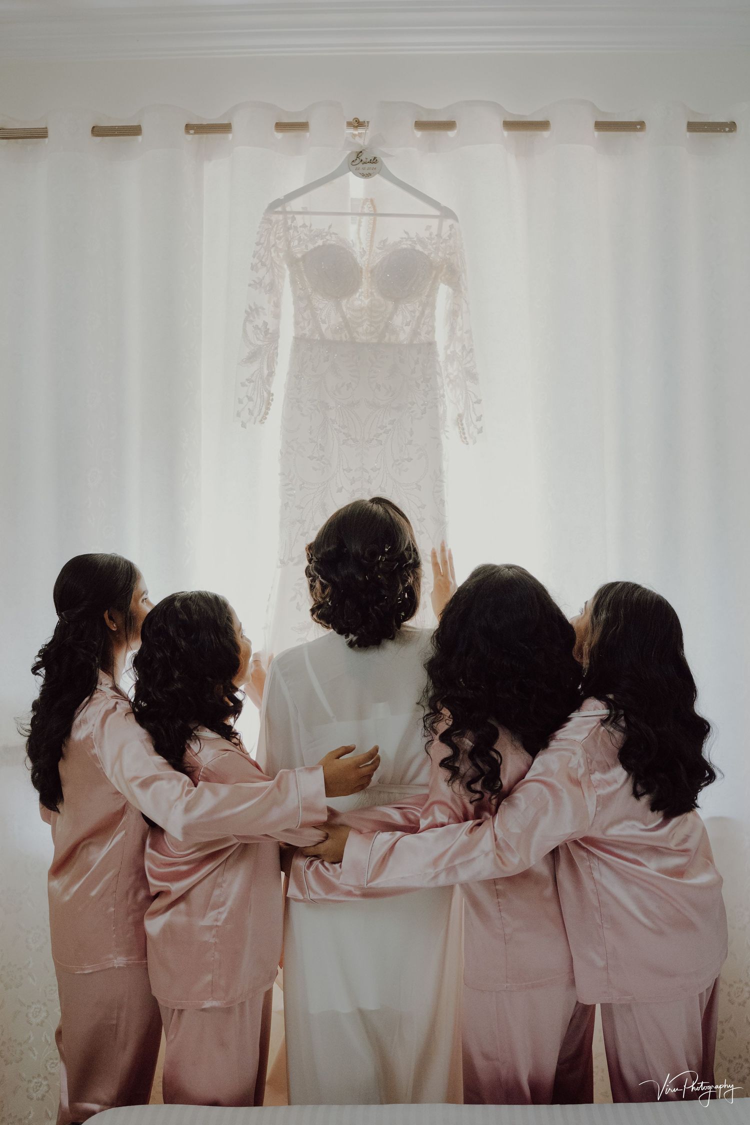 Wedding party in pink robes looking at hanging white lace bridal gown in front of white curtains.